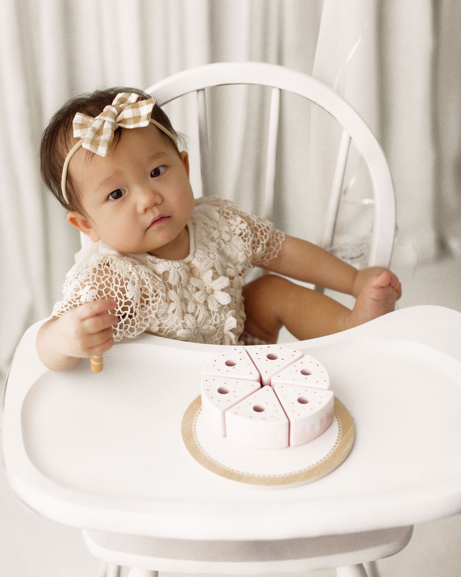 Baby in a lace dress and plaid hair bow sits in a white high chair beside a pink wooden toy birthday cake.