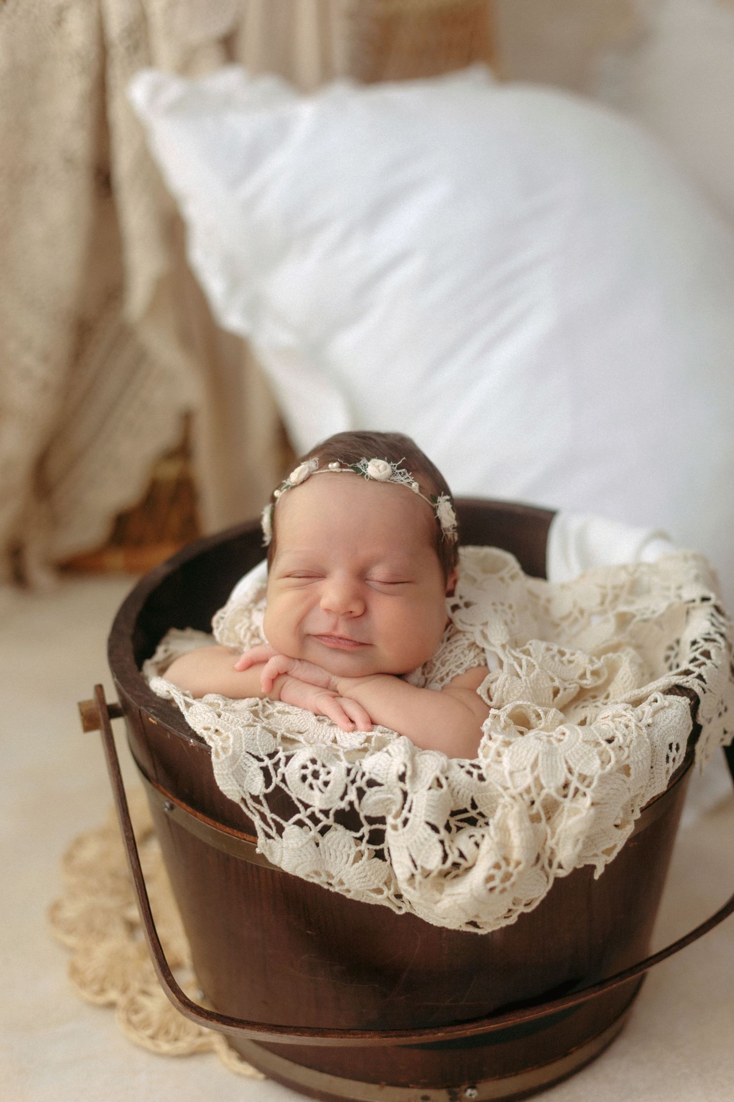 Sleeping newborn baby in wooden bucket with lace blanket and floral headband, soft white background.