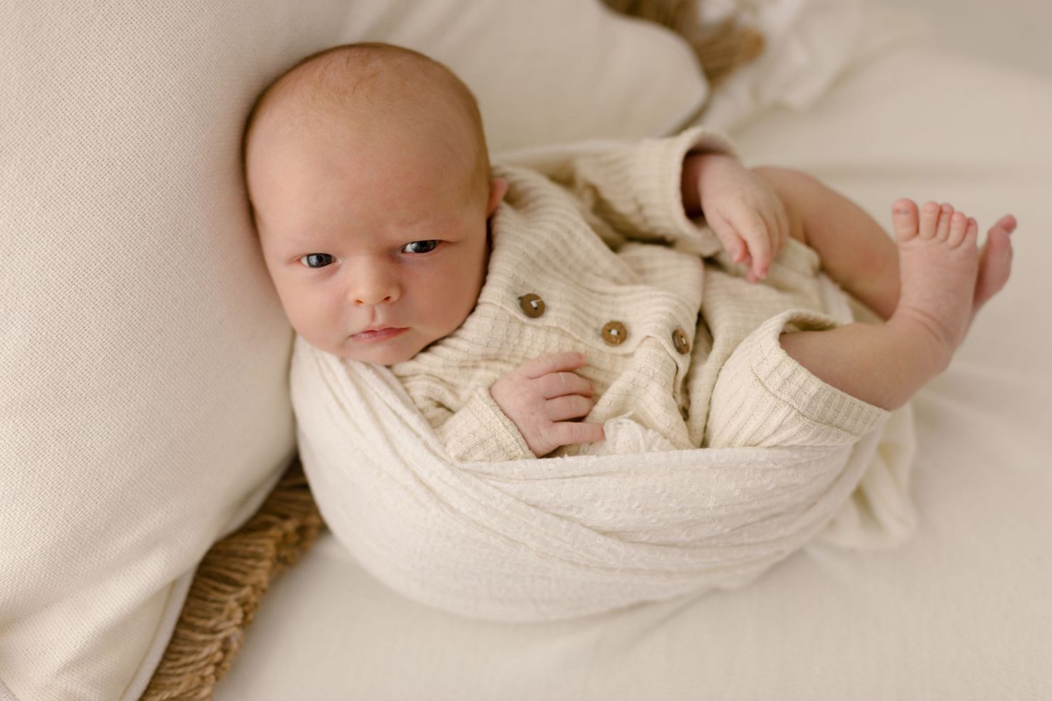 Newborn baby wrapped in cream knit blanket wearing beige button cardigan, lying on white soft bedding.