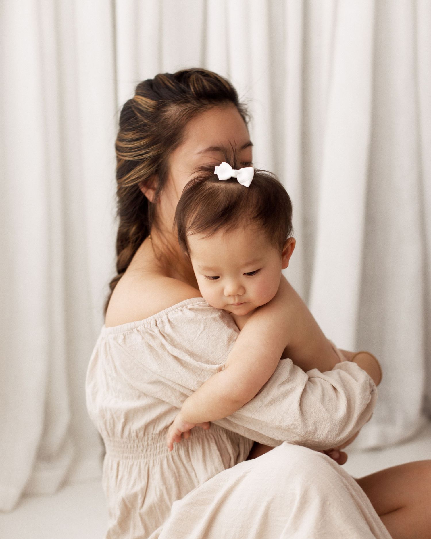 A mother in a cream off-the-shoulder dress cradles her baby, who wears a white bow, against soft white curtains in a bright studio.