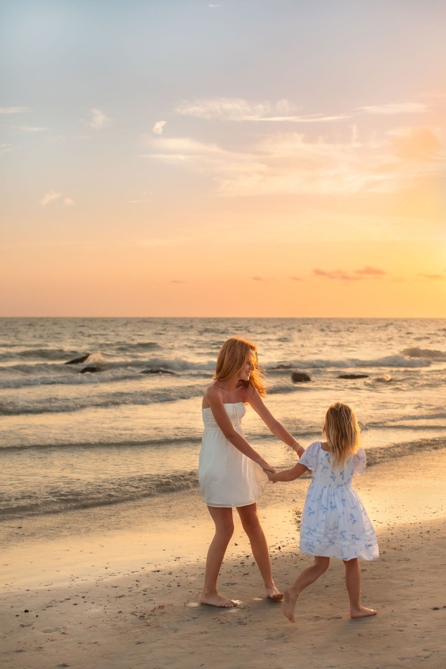 Mother and daughter in white dresses holding hands dancing on a sandy beach at golden sunset.