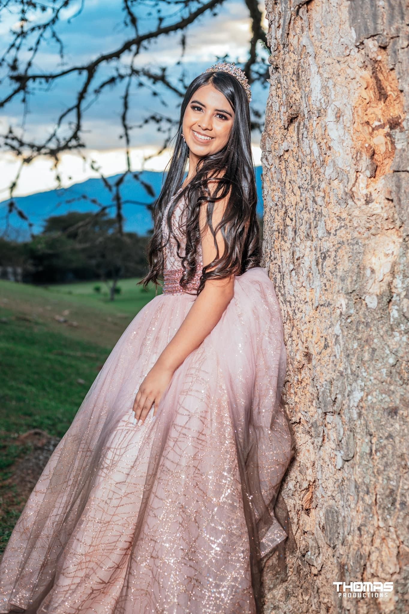A woman in a pink gown poses against a tree trunk during a golden hour photoshoot.