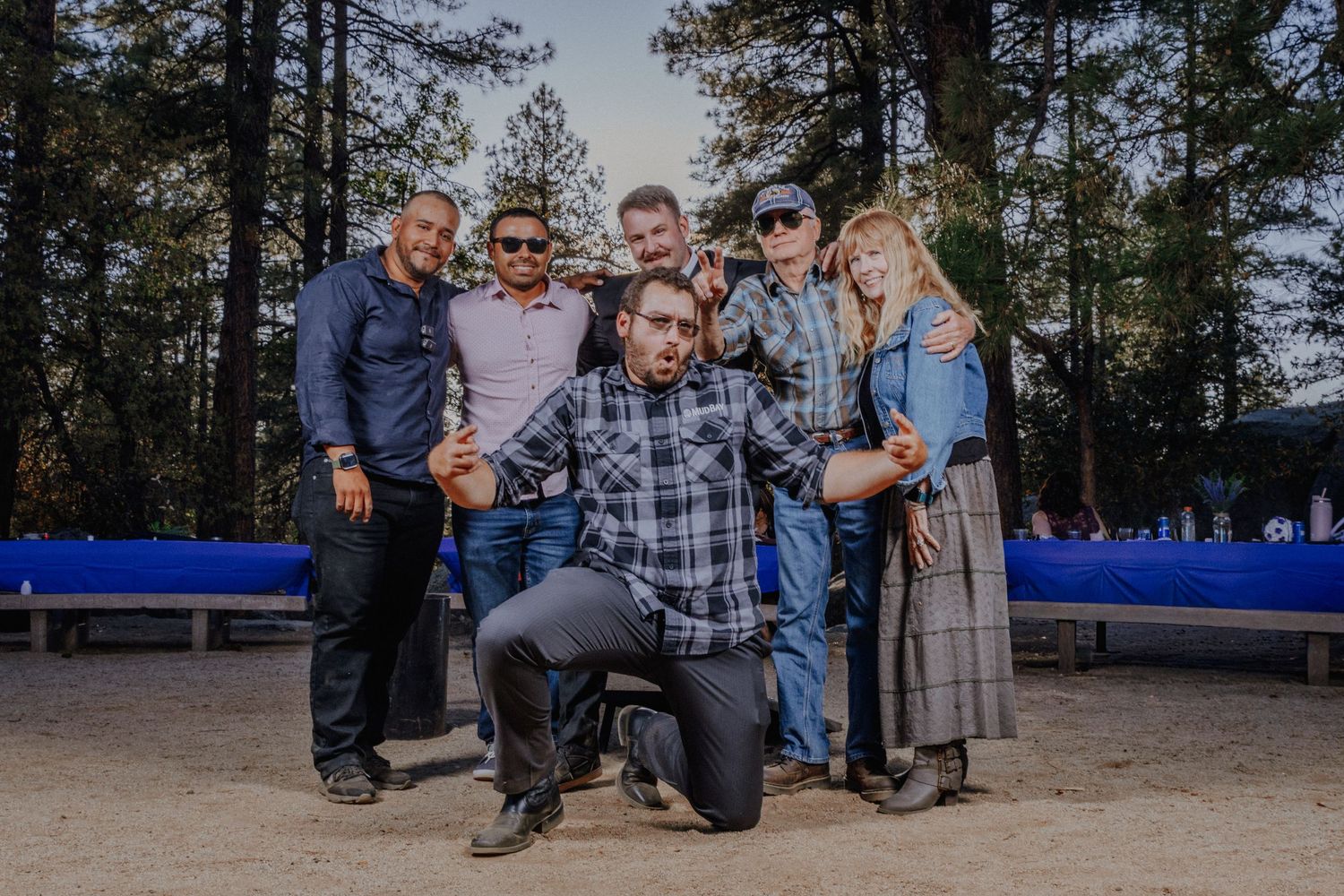 Group photo of wedding party on wooden benches in outdoor forest setting.