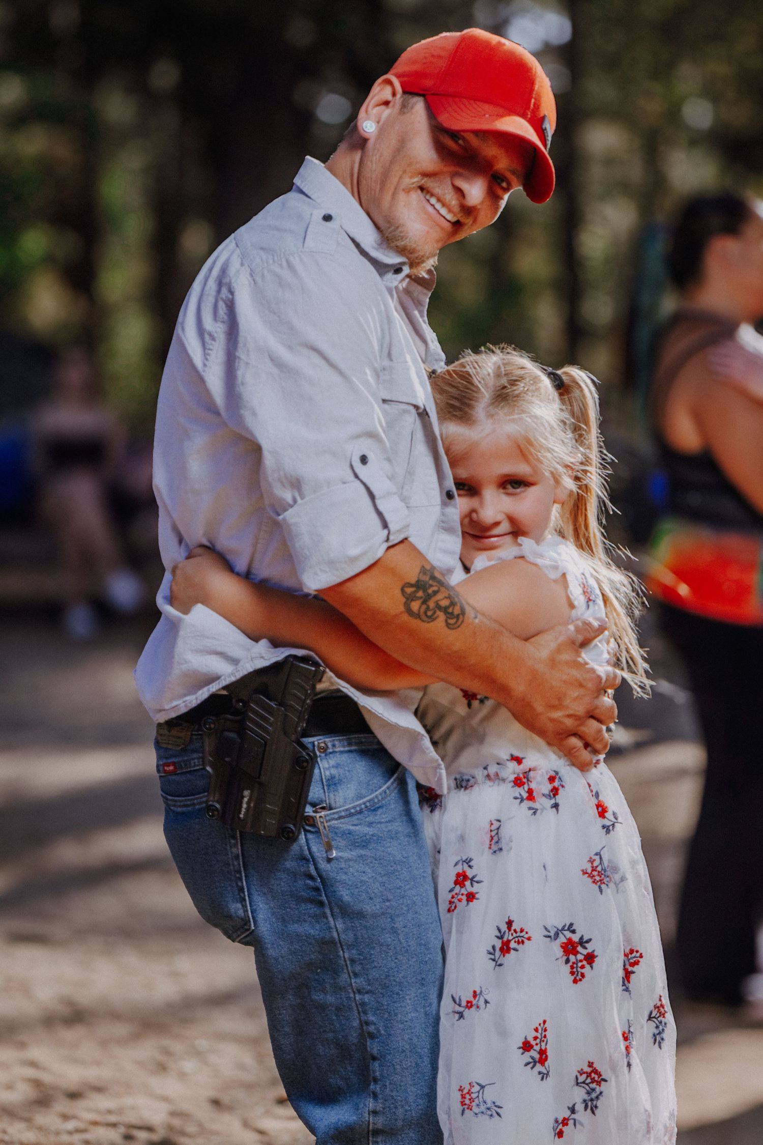 Couple embracing outdoors with woman wearing floral dress and man in red baseball cap and jeans.