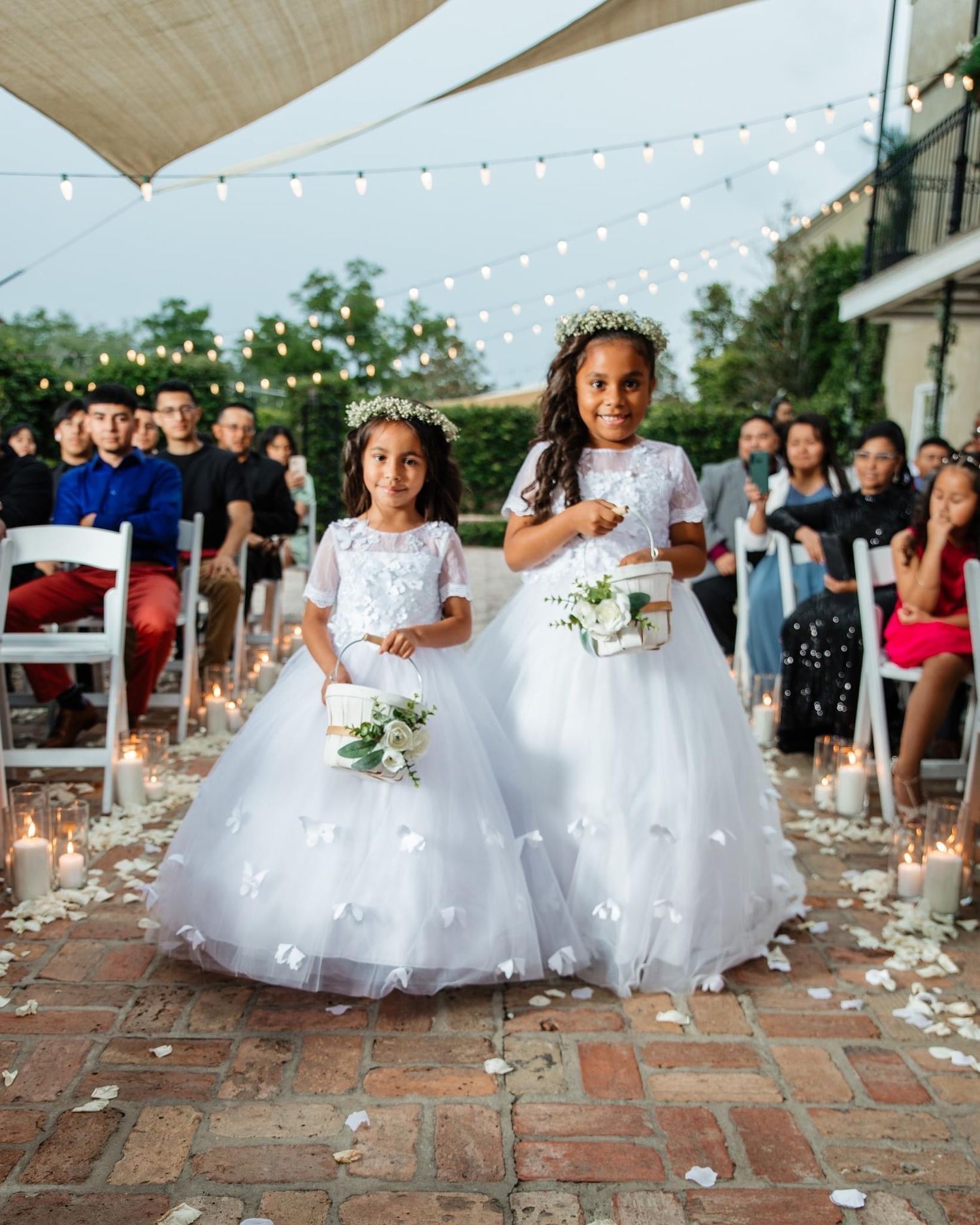 Young flower girls in white dresses walk down a brick path under string lights.