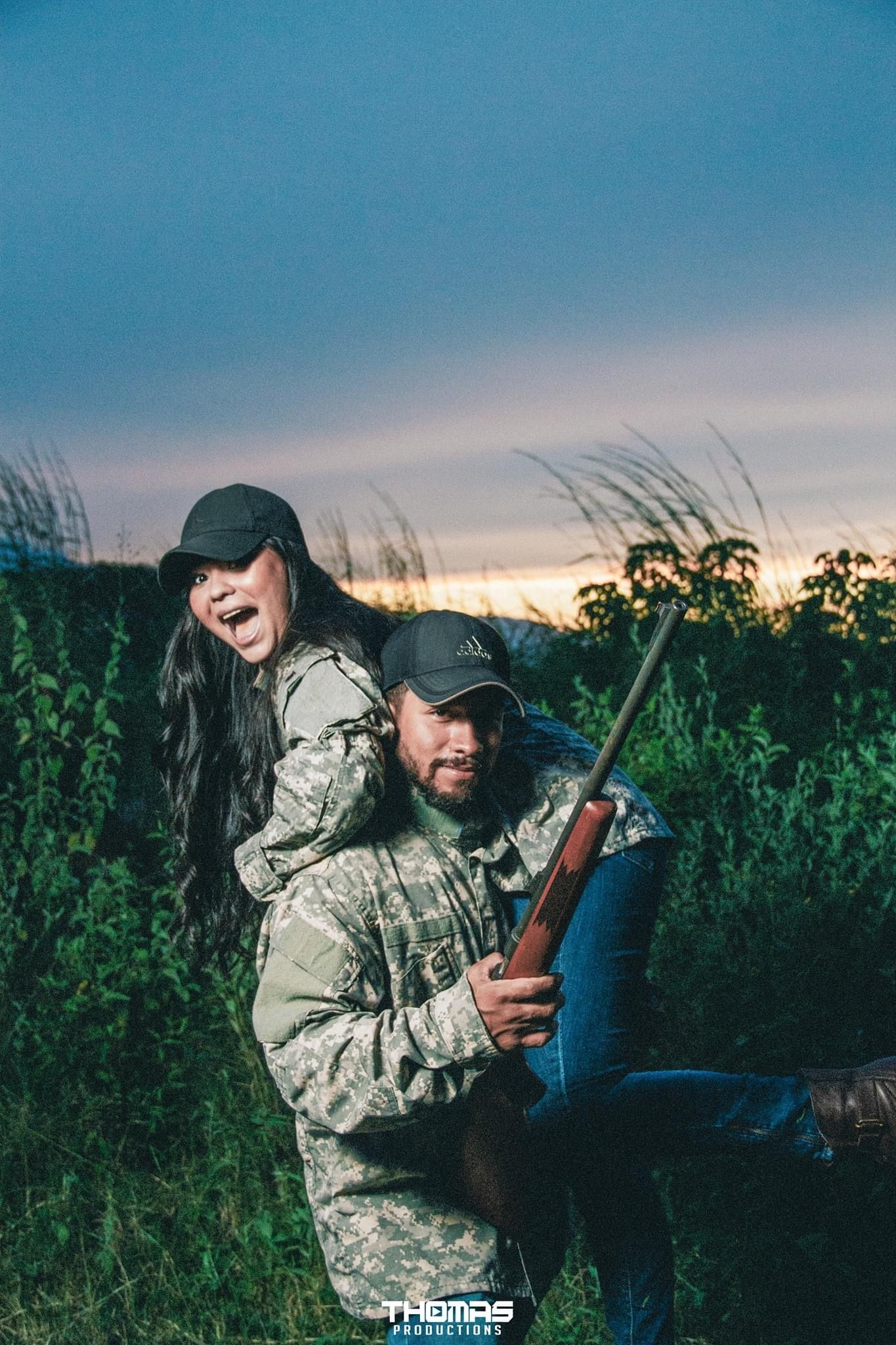 A person in camouflage gear poses with hunting equipment in a field at dusk.