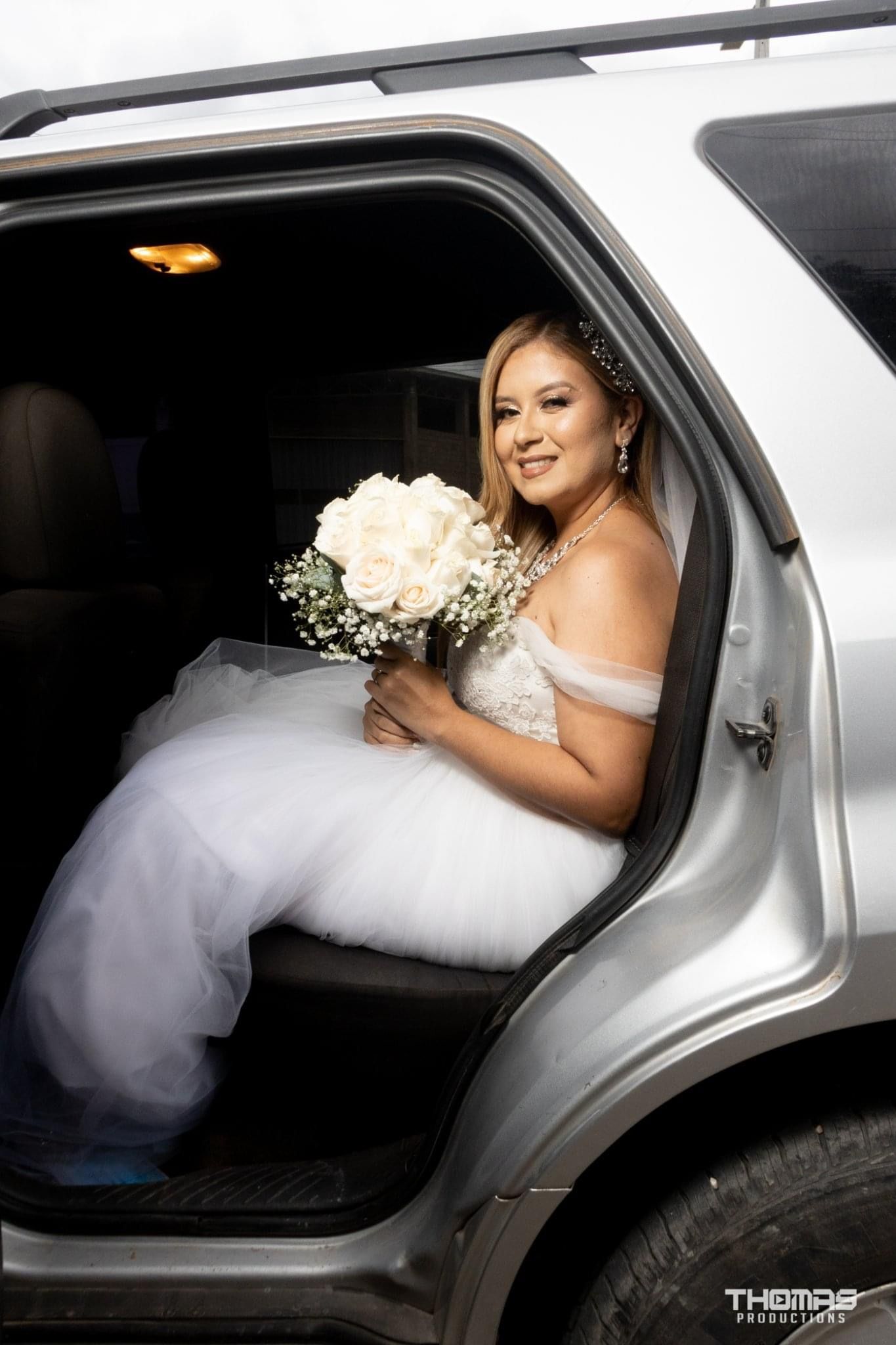 A bride in a white gown poses elegantly in the backseat of a luxury car.