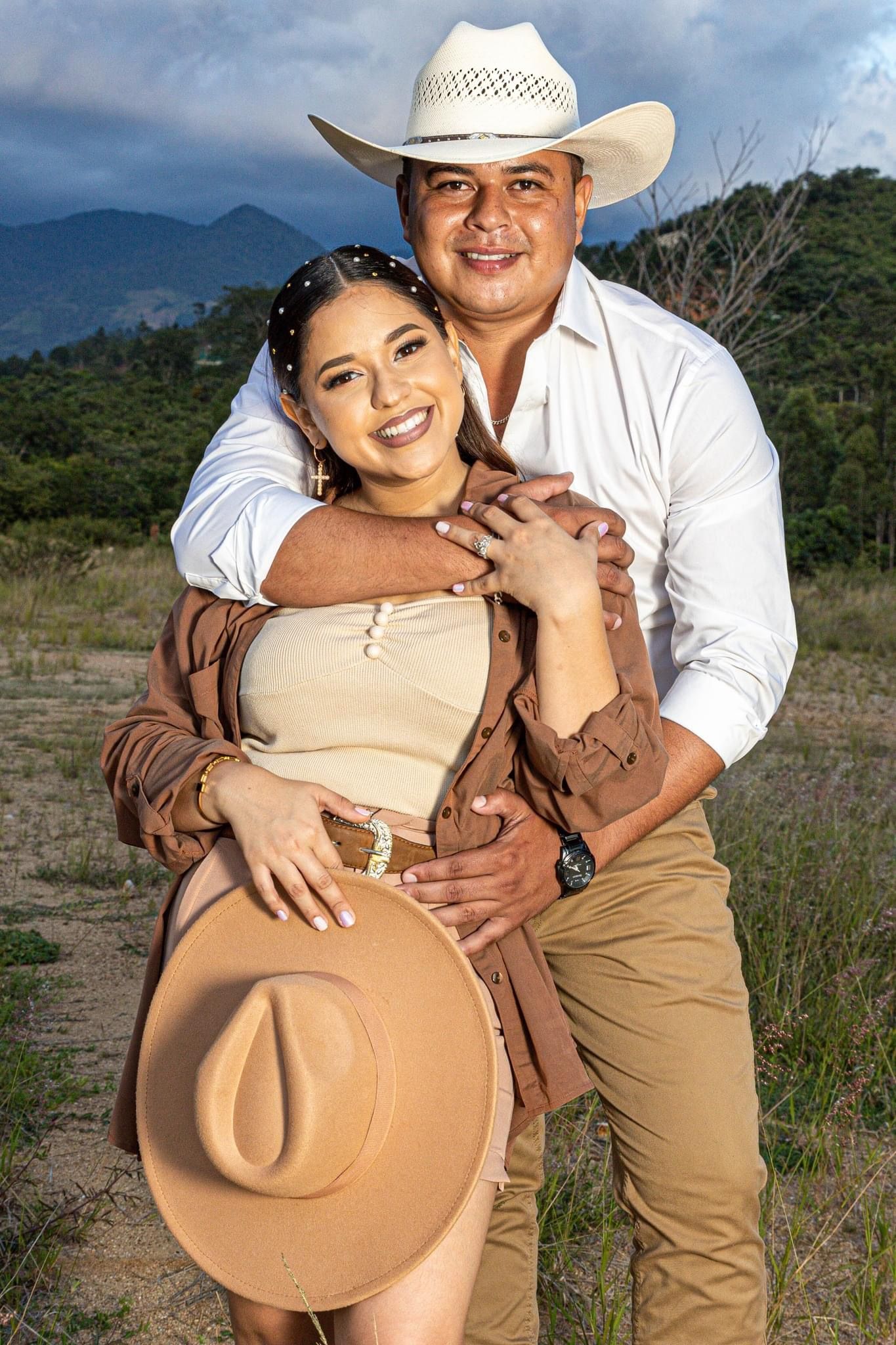 A couple poses for engagement photos in a rustic countryside setting with cowboy hats.