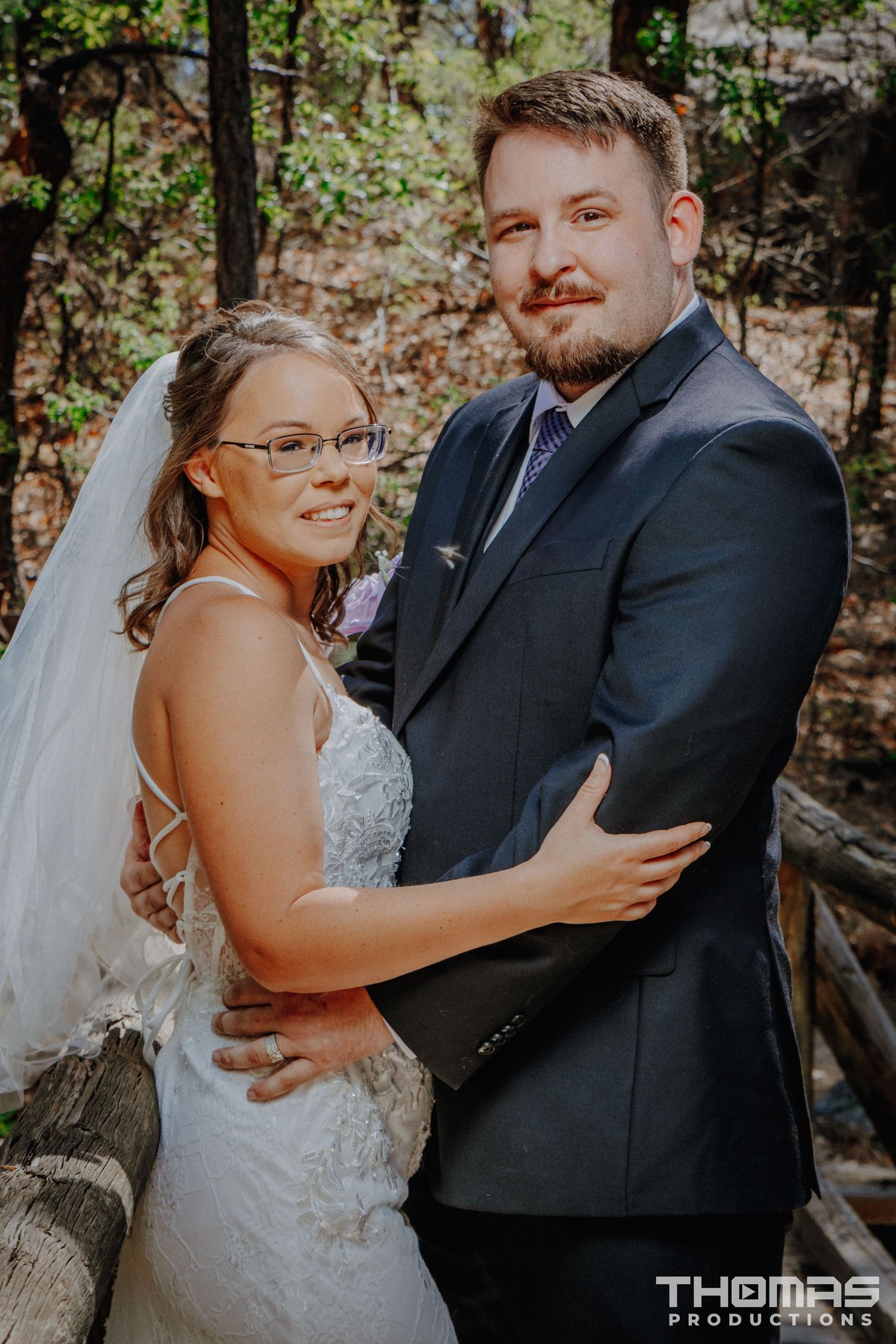 A bride in a lace dress and groom in a dark suit embrace on a wooden bridge in the forest.