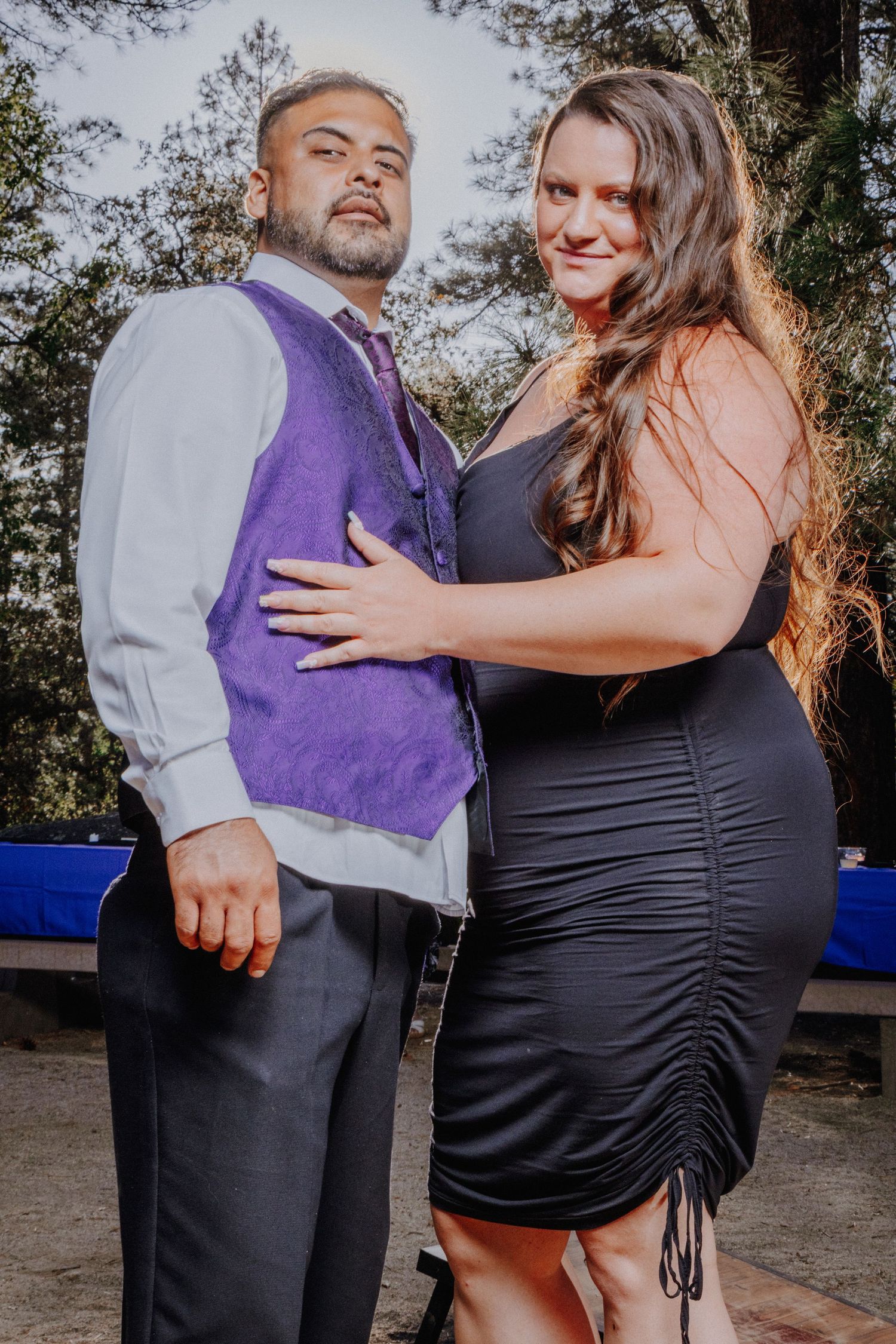 Couple in formal attire embracing during outdoor photo session in wooded setting.