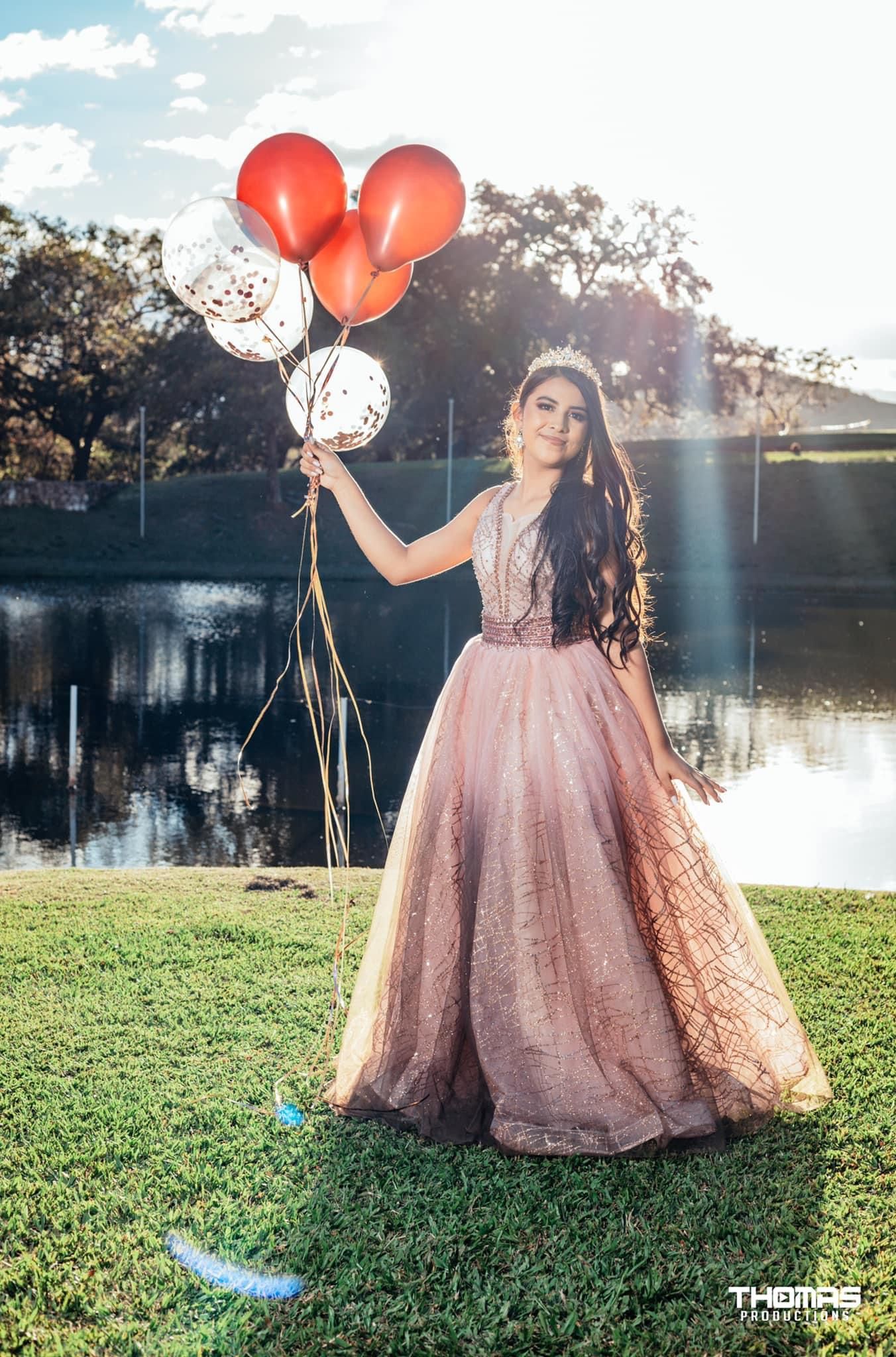 A person in a pink sparkly ballgown poses with red balloons by a lake during sunset.