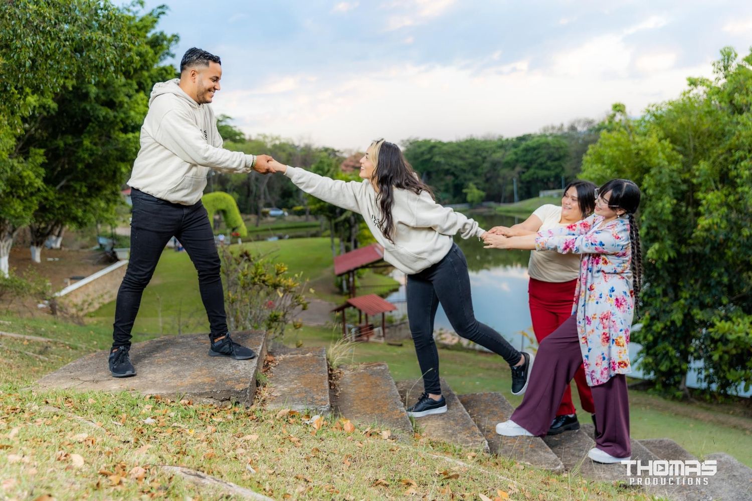 Playful family photo session with group holding hands in park setting near water.
