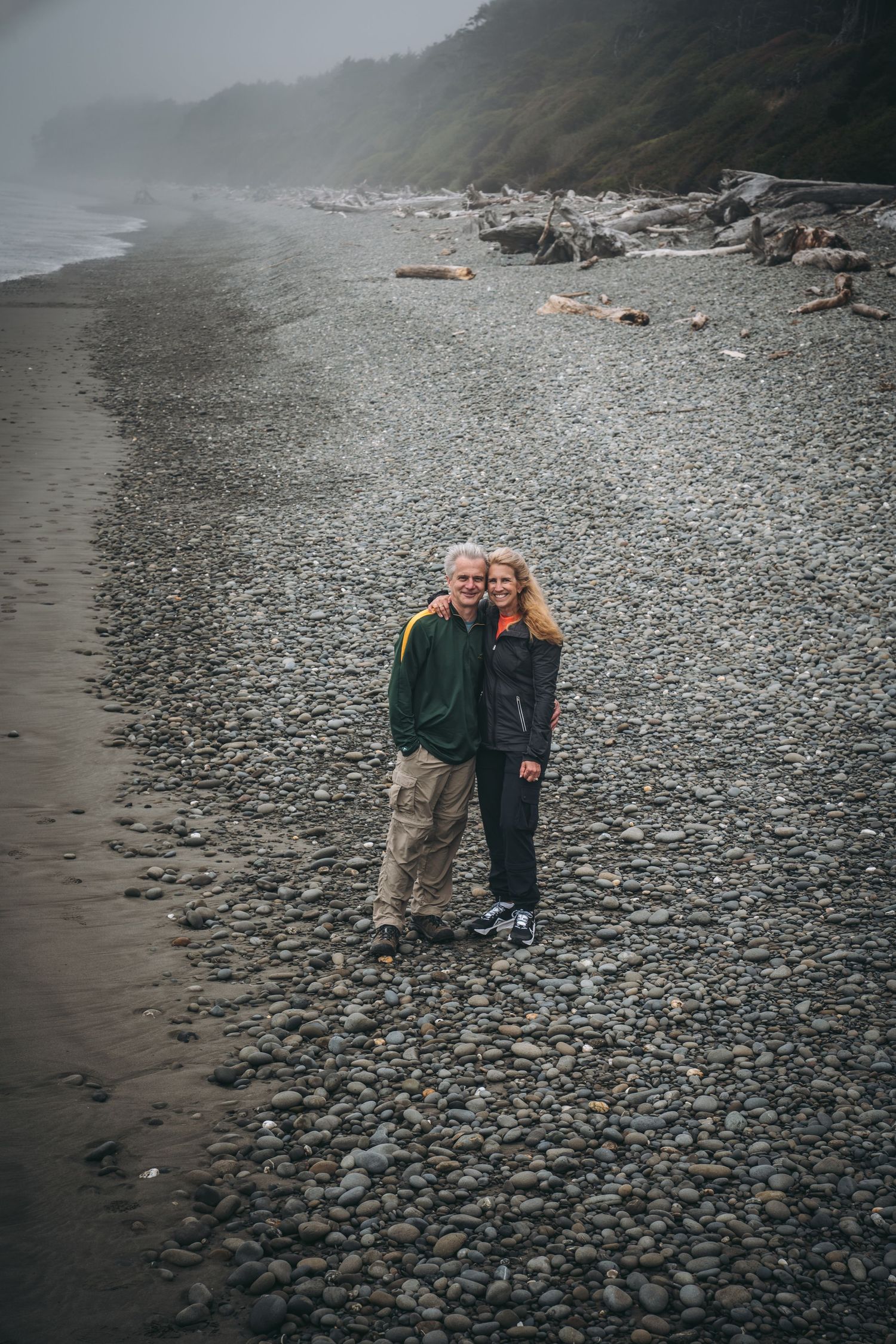 Smiling couple embracing on a rocky foggy beach with driftwood in the background.