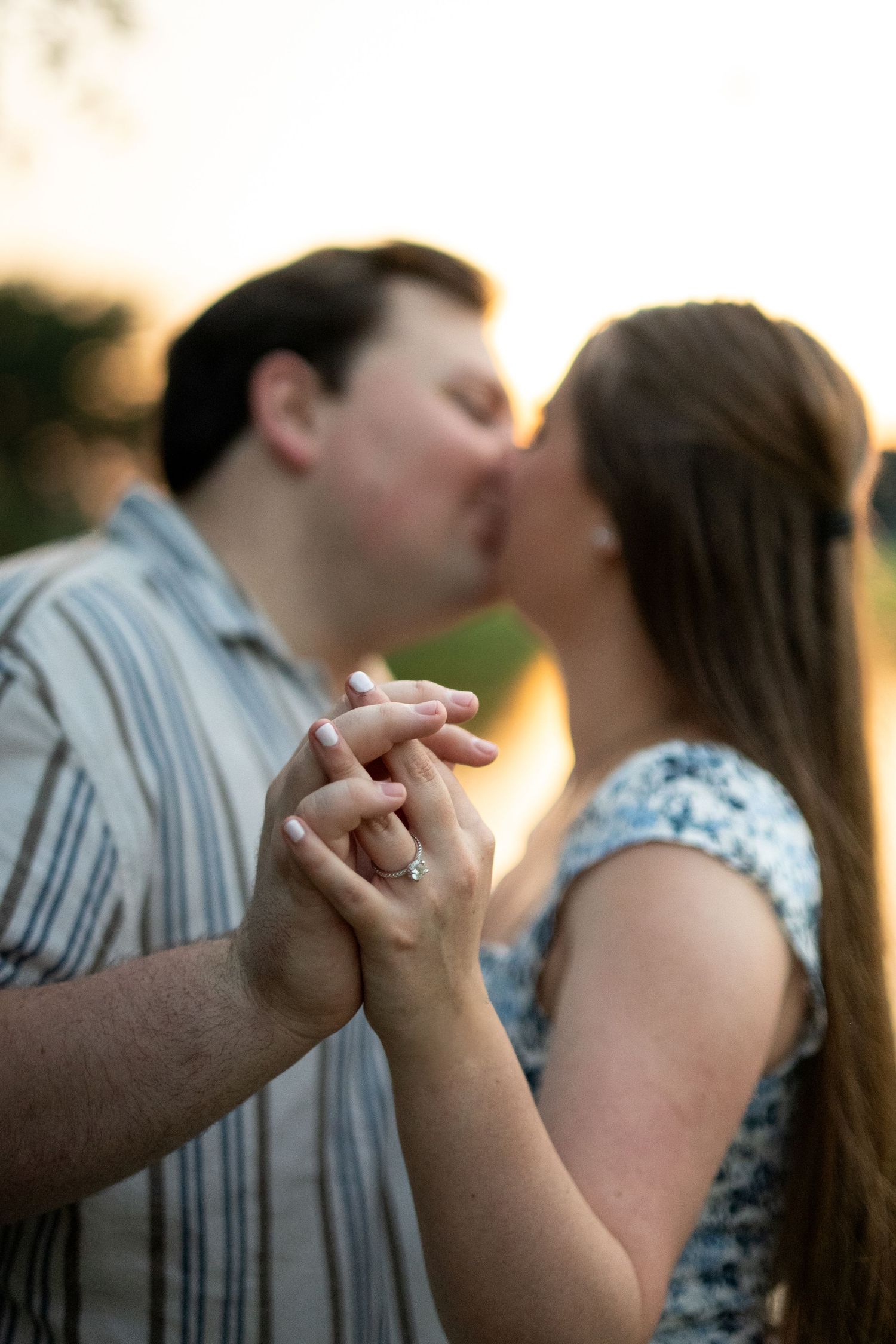 Couple kissing at sunset while holding hands, engagement ring visible on woman's finger.