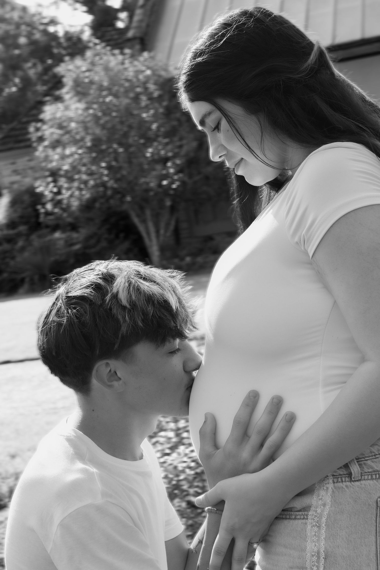 Black and white photo of a man kissing a pregnant woman's belly outdoors, a tender maternity moment.