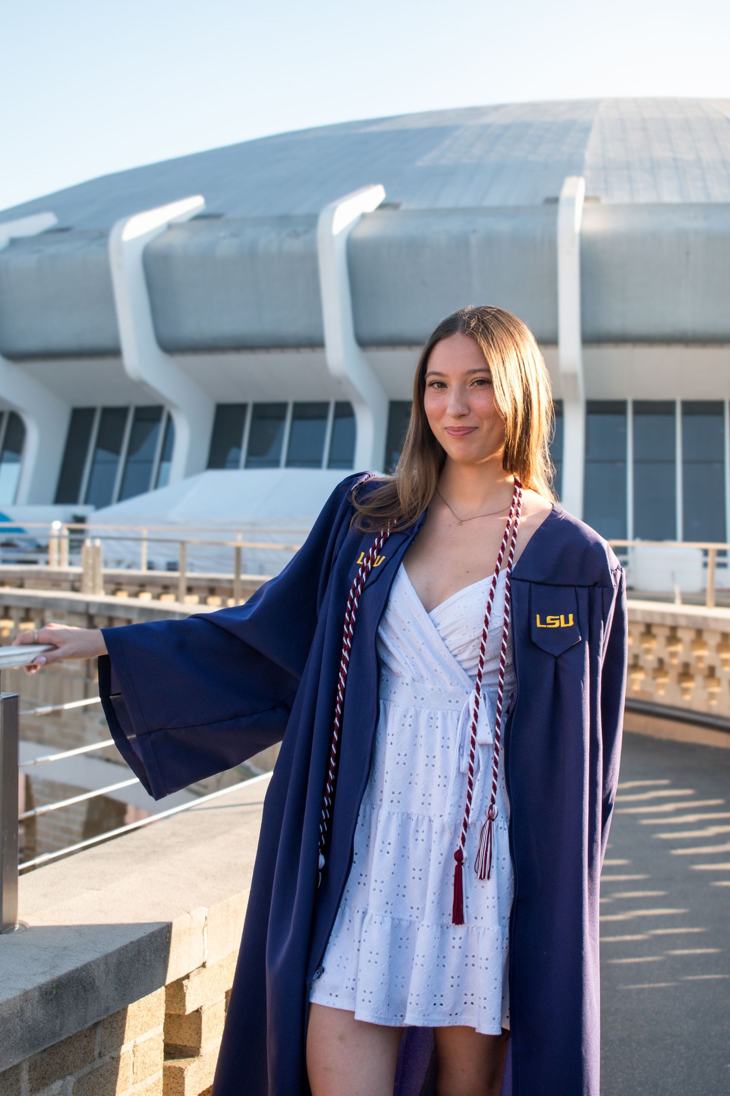 Female LSU graduate in navy gown with red and white cords poses near modern stadium.