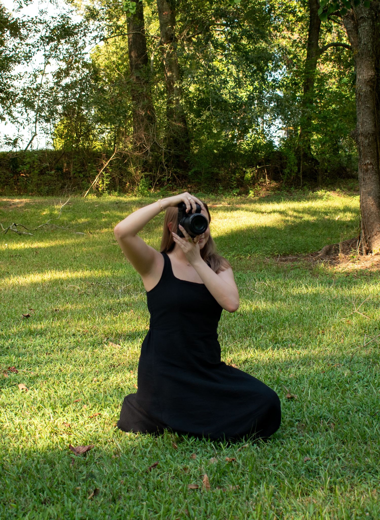 Woman in black dress kneeling on grass, photographing with DSLR camera, surrounded by lush green trees outdoors.