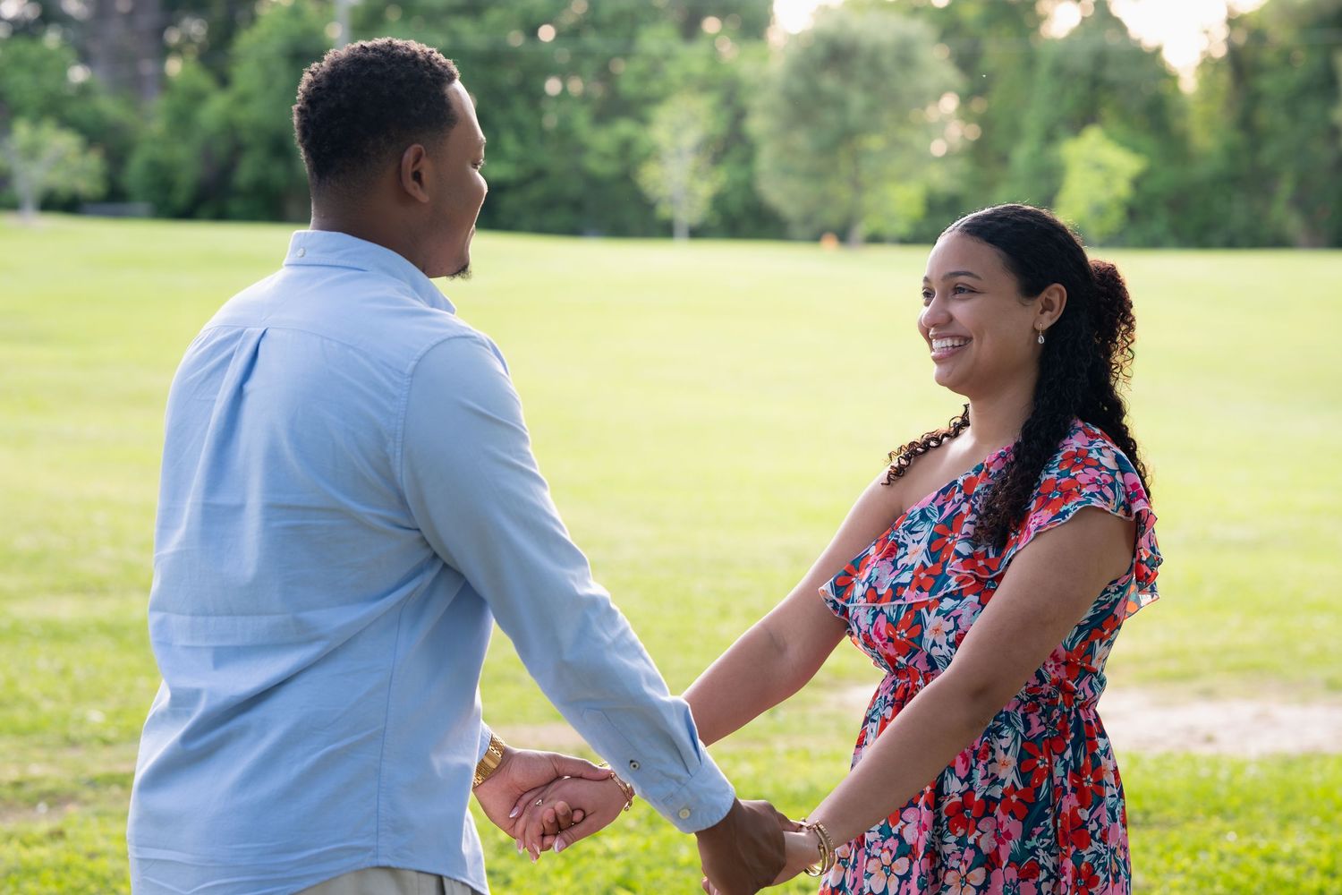Couple holding hands and smiling at each other in a sunny green park, wearing casual colorful outfits.