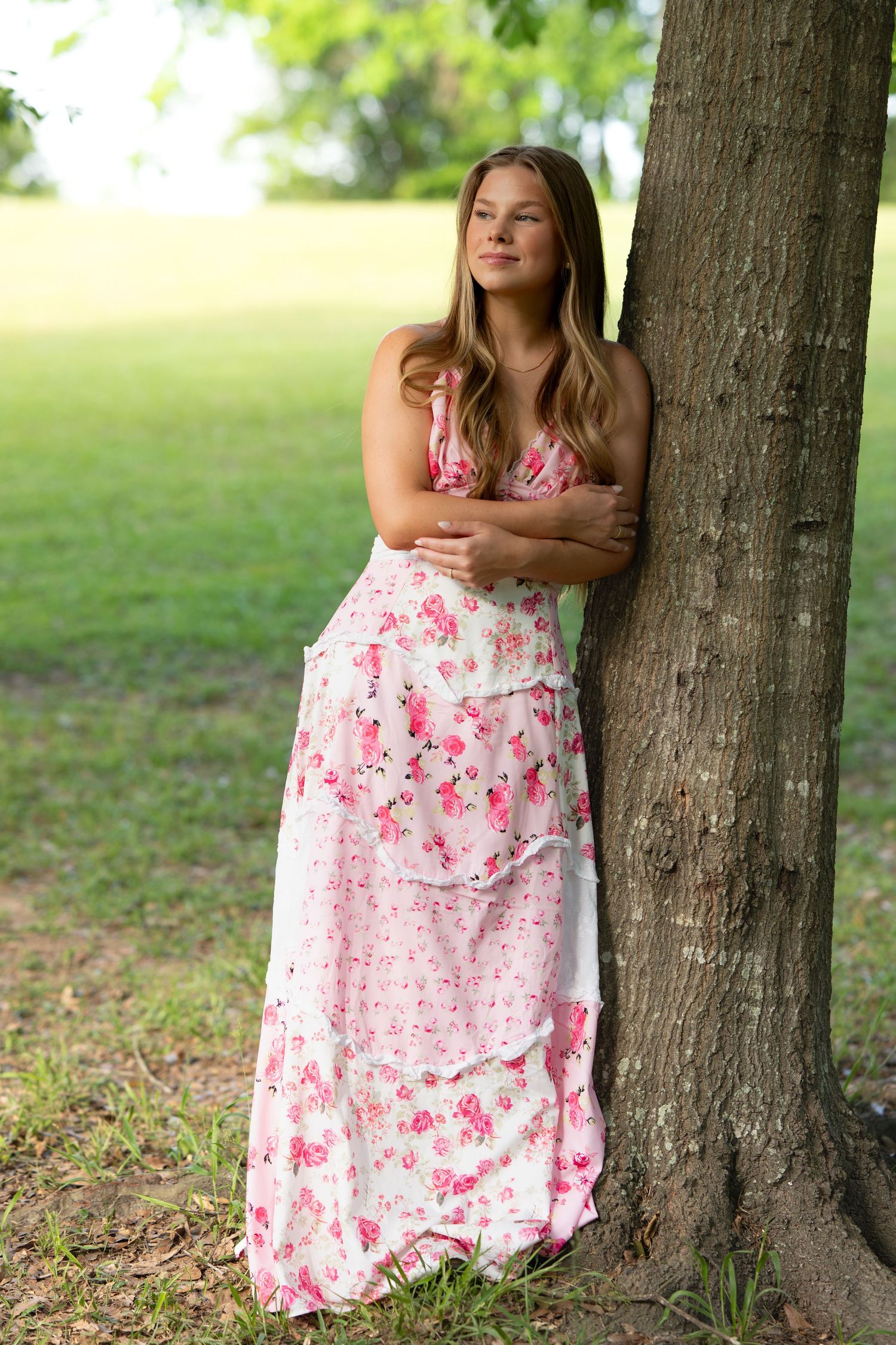 Young woman in pink floral maxi dress leaning against tree trunk in lush green park setting.