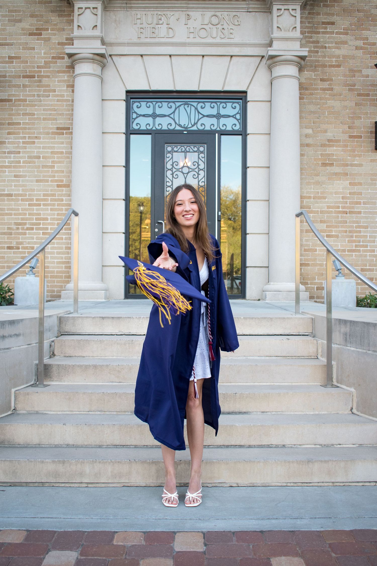 Young woman in navy blue graduation gown and cap smiles outside a brick university building on graduation day.