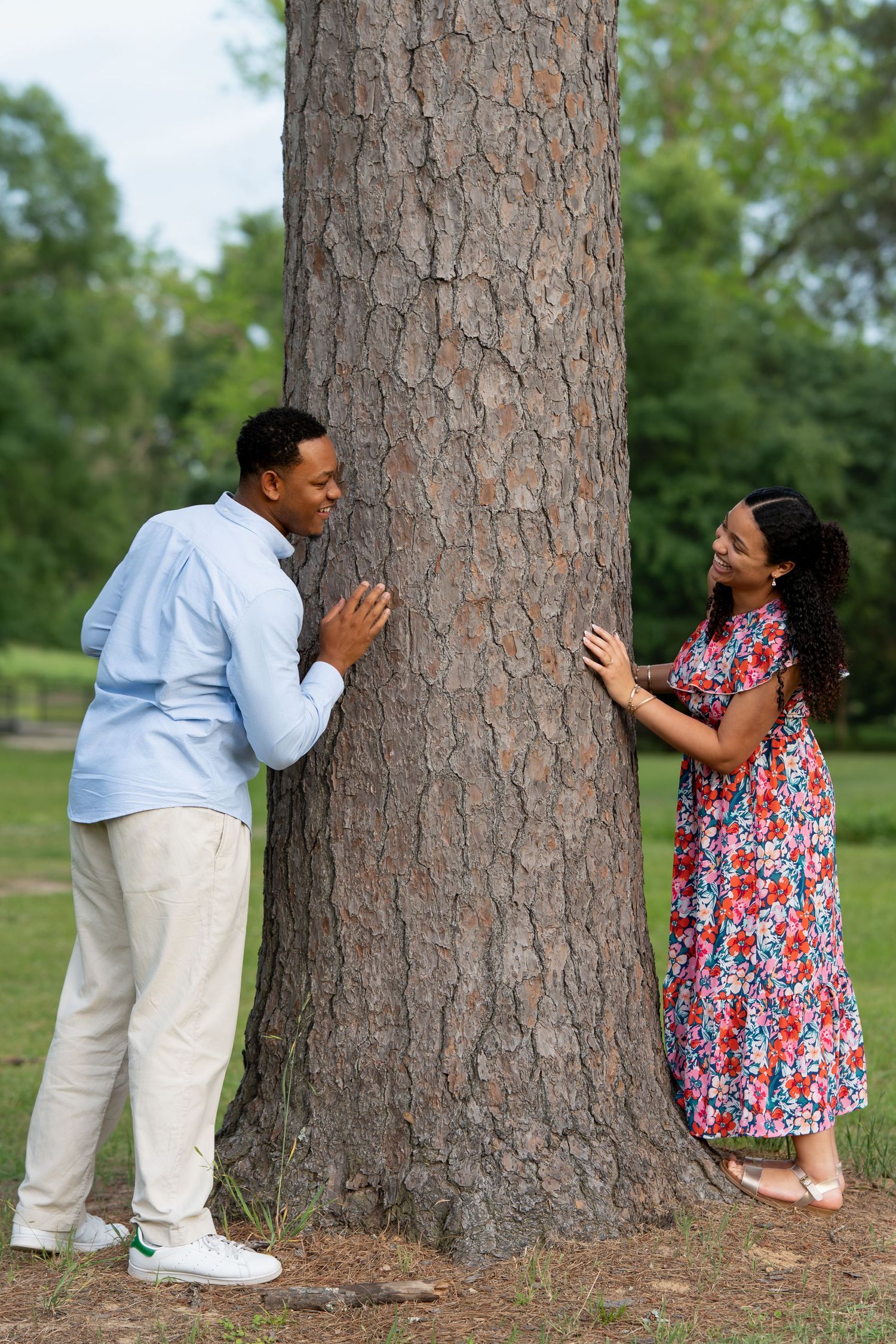Playful couple peeking at each other from opposite sides of a large tree trunk in a park.