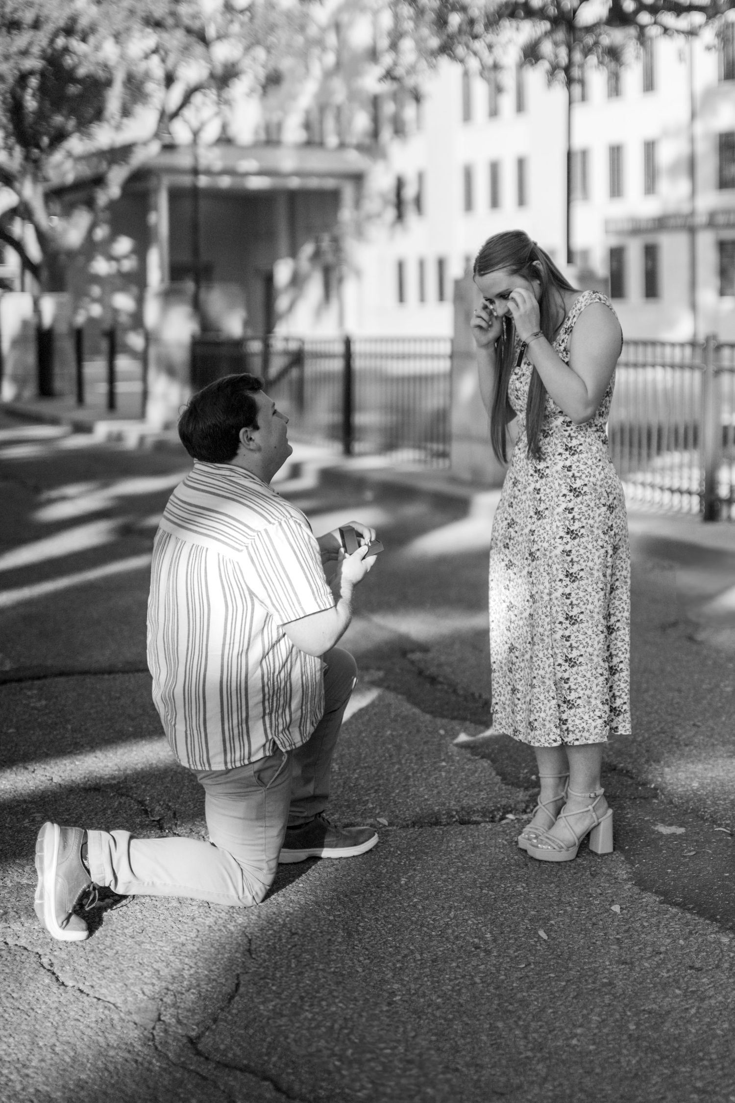 Black and white photo of a man on one knee proposing to an emotional woman on a sunny outdoor path.