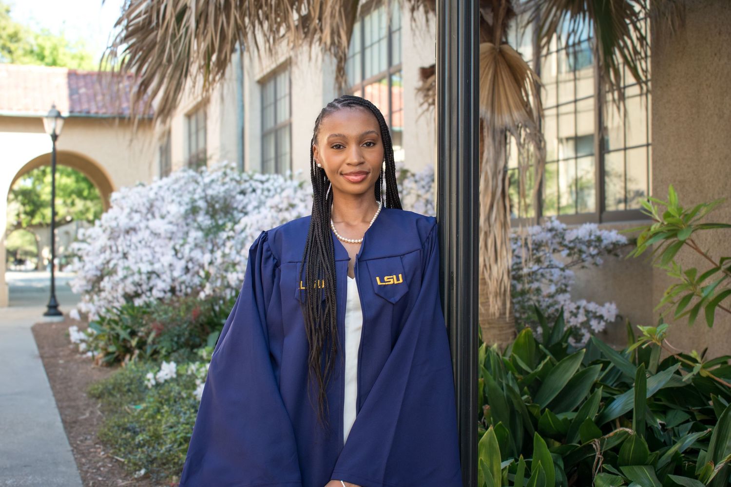 Young woman in LSU graduation gown smiling outdoors near palm trees and flowering bushes on campus.