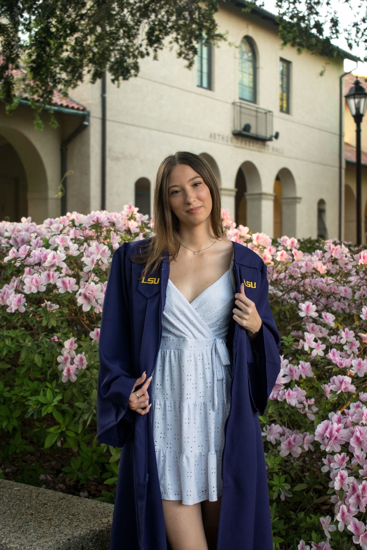 LSU graduate poses in navy cap and gown over white dress, surrounded by pink azaleas on campus.