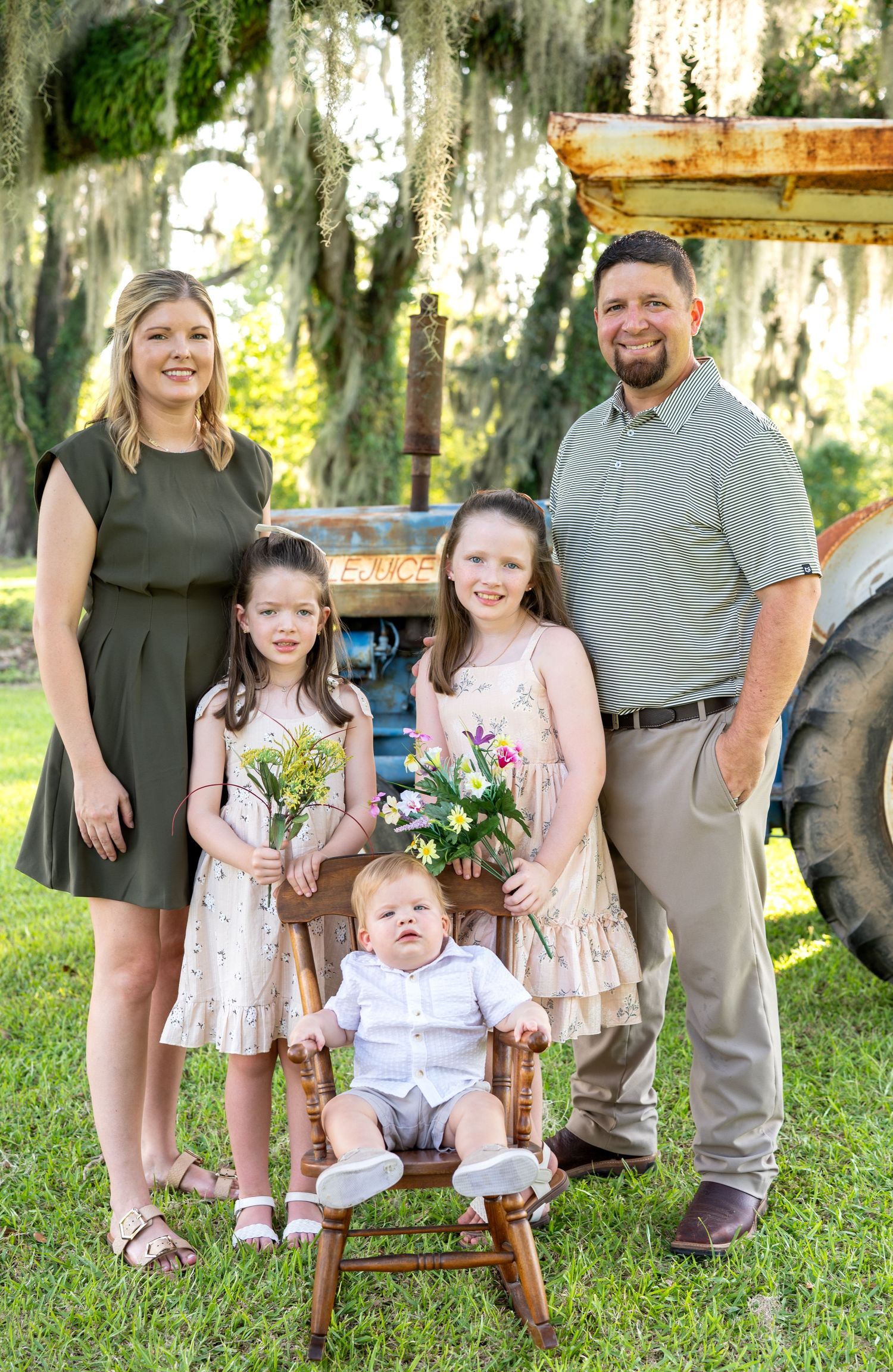 Family portrait of two parents, two girls holding wildflowers and a baby on a wooden rocking chair beside a vintage tractor under oak trees.