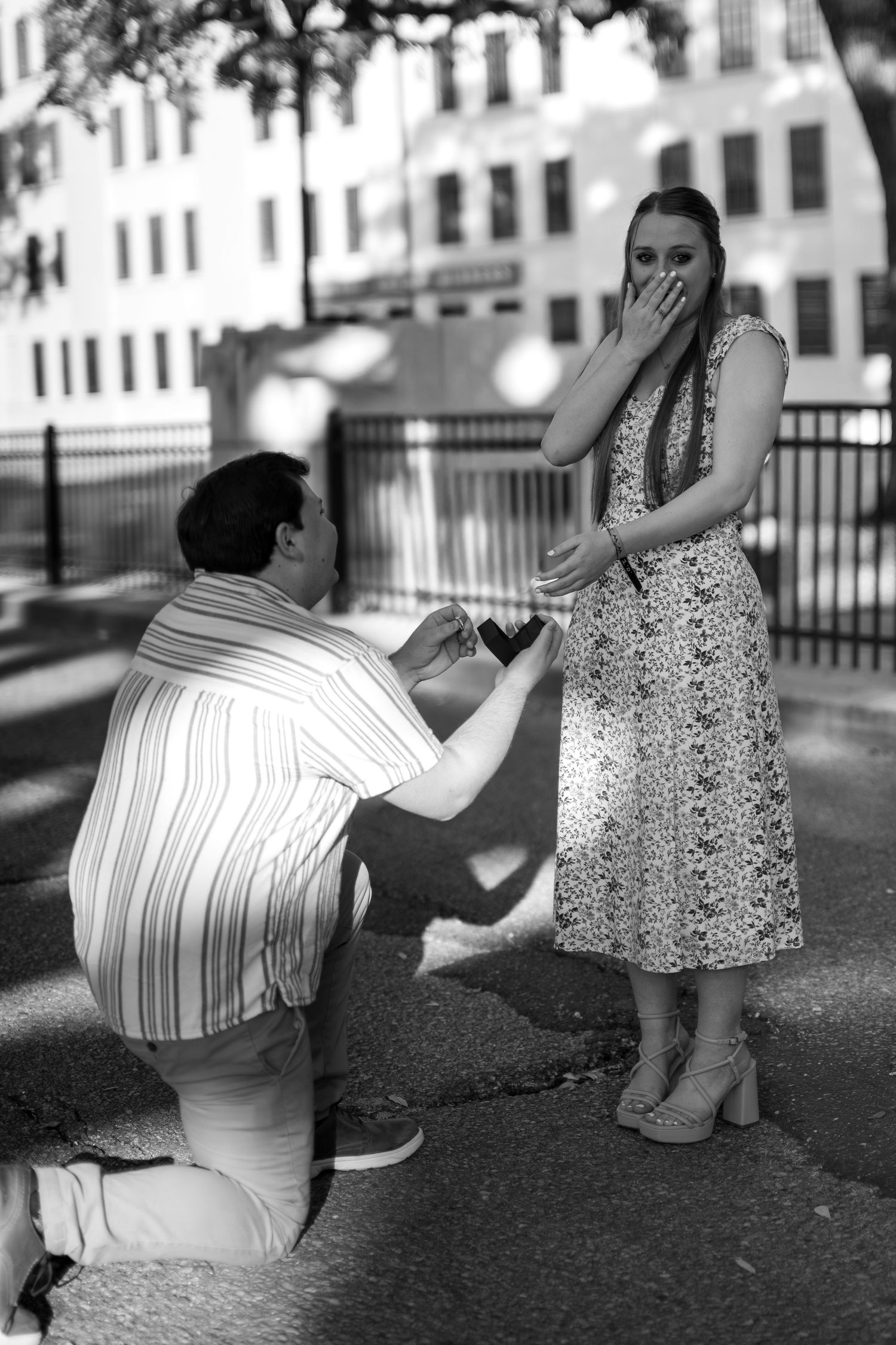 Black and white photo of a man on one knee proposing to a surprised woman in a floral dress outdoors.