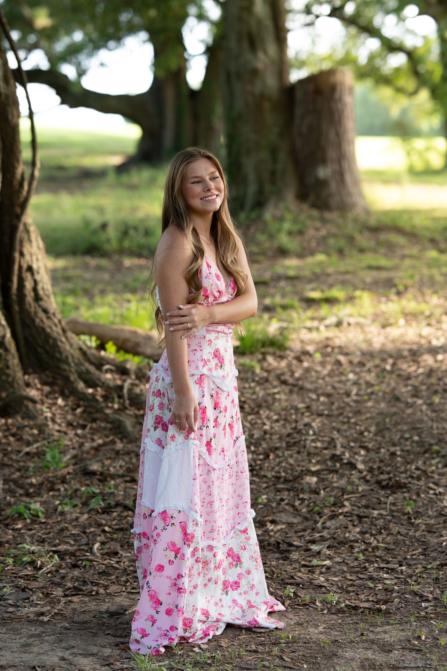 Young woman in pink floral maxi dress smiling outdoors near large oak trees in a garden setting.