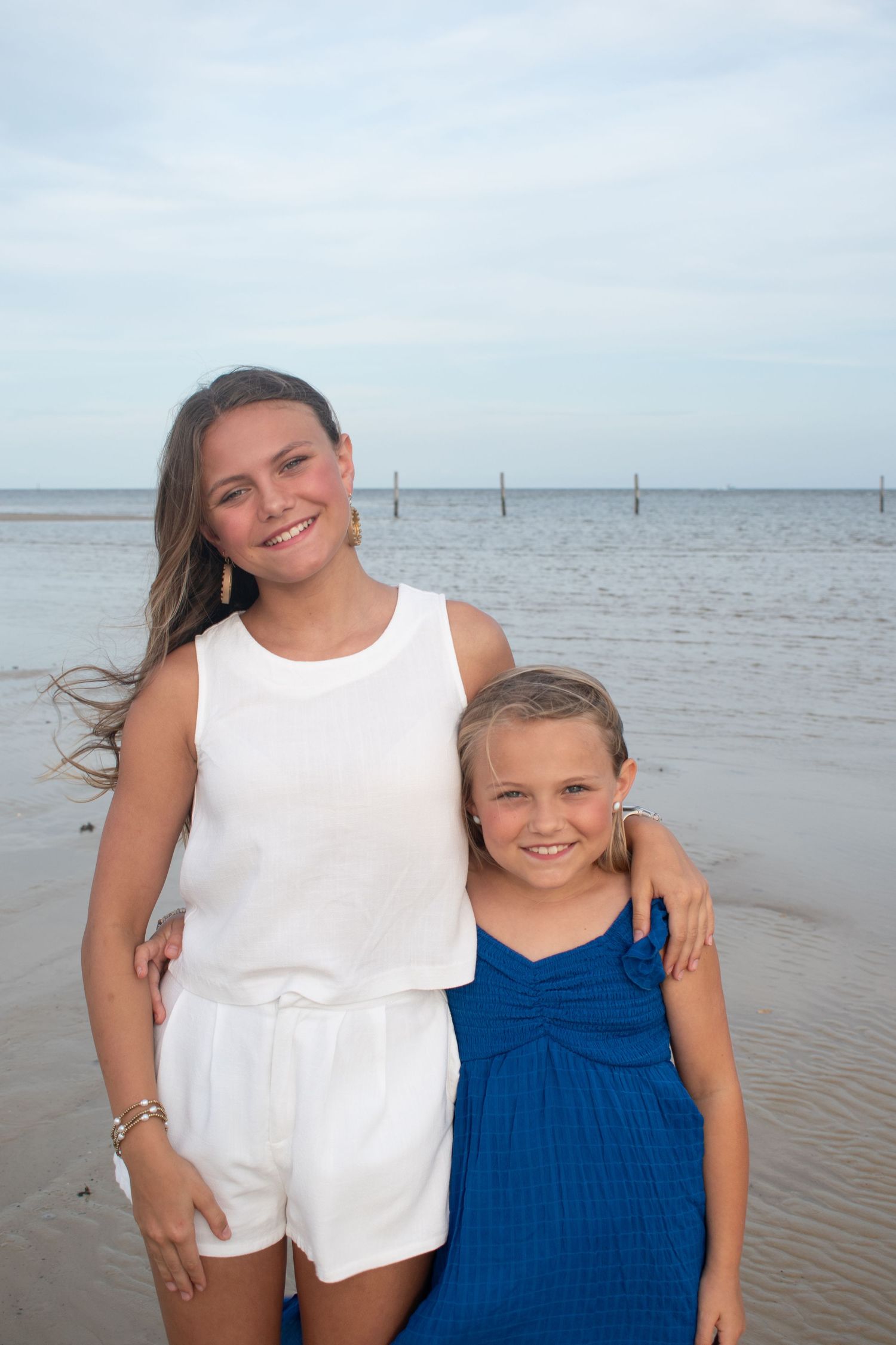 Two sisters smile together on a beach, one in white and one in a blue dress.
