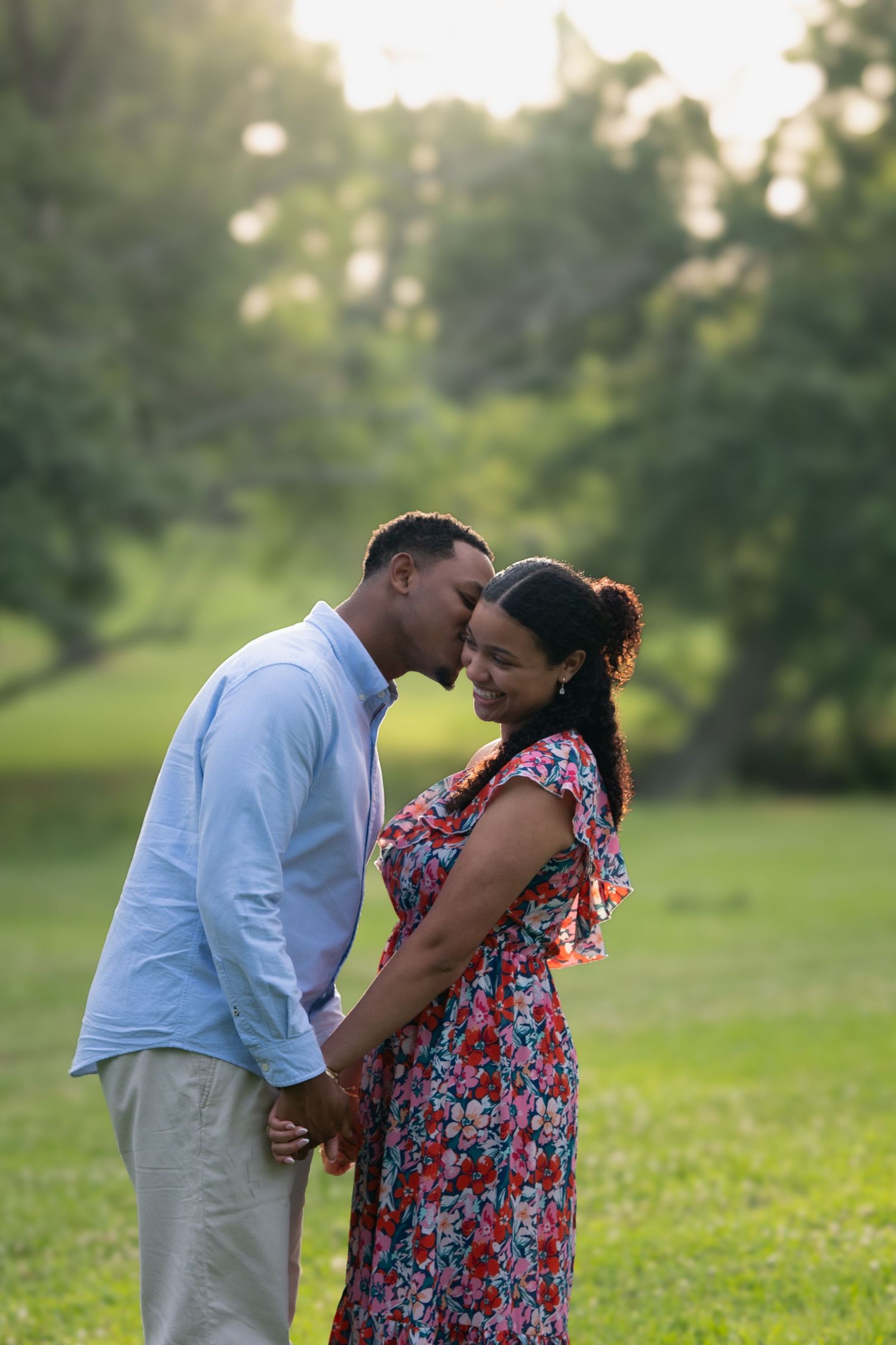 Tender moment as man kisses womans forehead while holding hands in a sunlit green park.