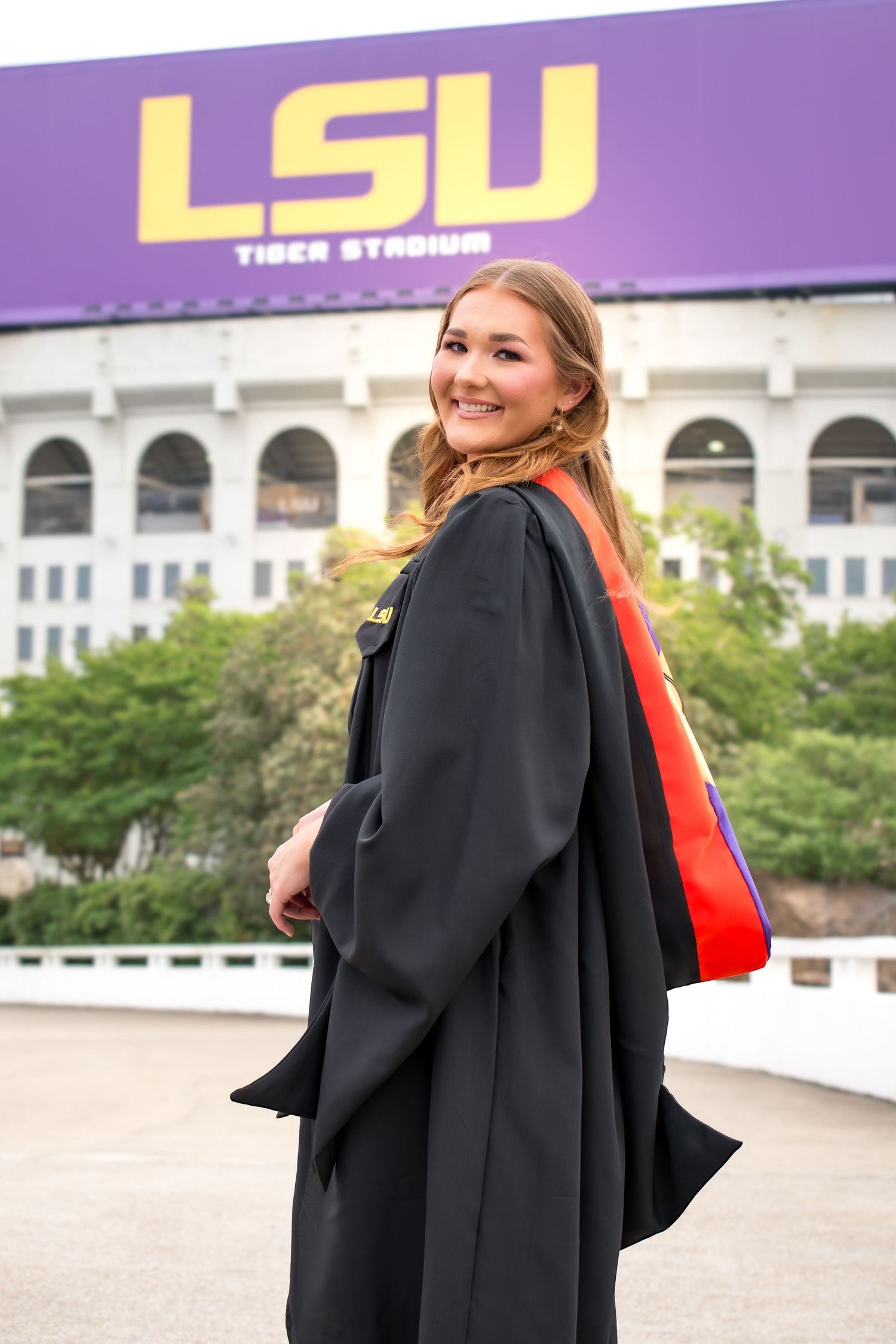 Female LSU graduate in black doctoral gown smiling over shoulder in front of LSU Tiger Stadium sign.
