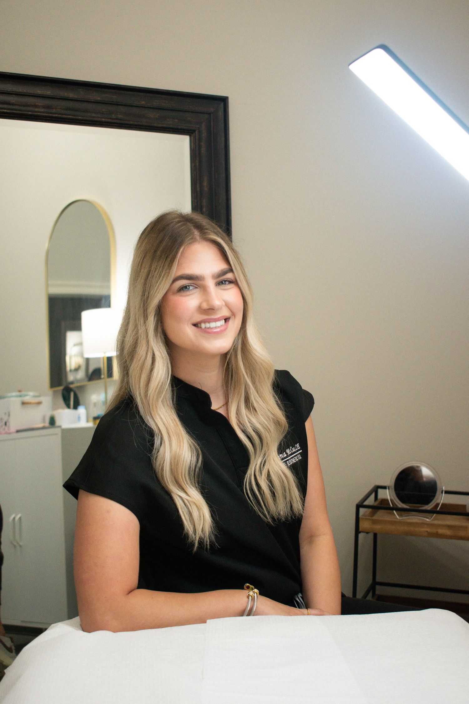A smiling blonde esthetician in black scrubs sits at a treatment table in a modern skincare studio.
