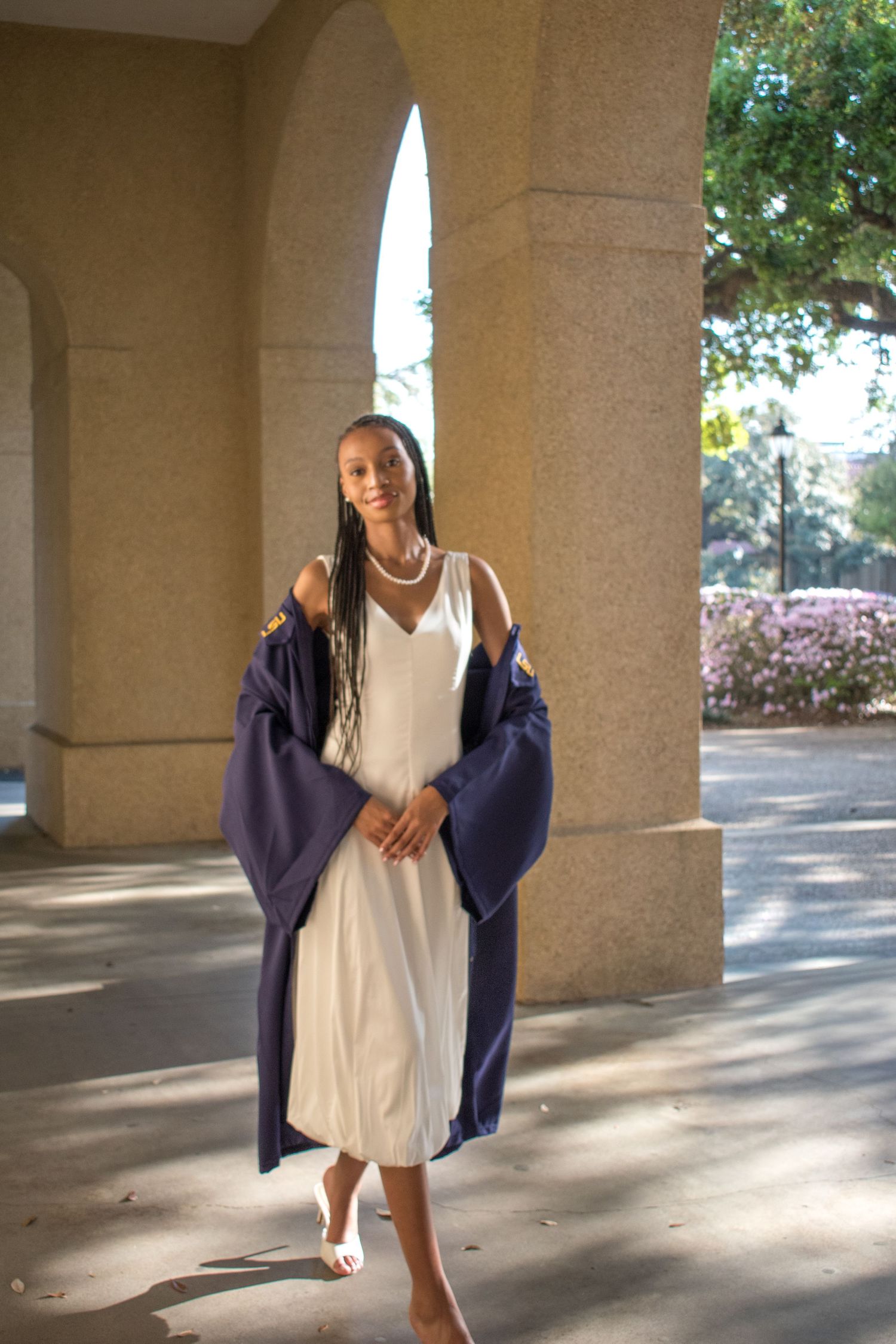 Young woman in white dress holding navy graduation gown, posing between stone columns on a sunny campus.