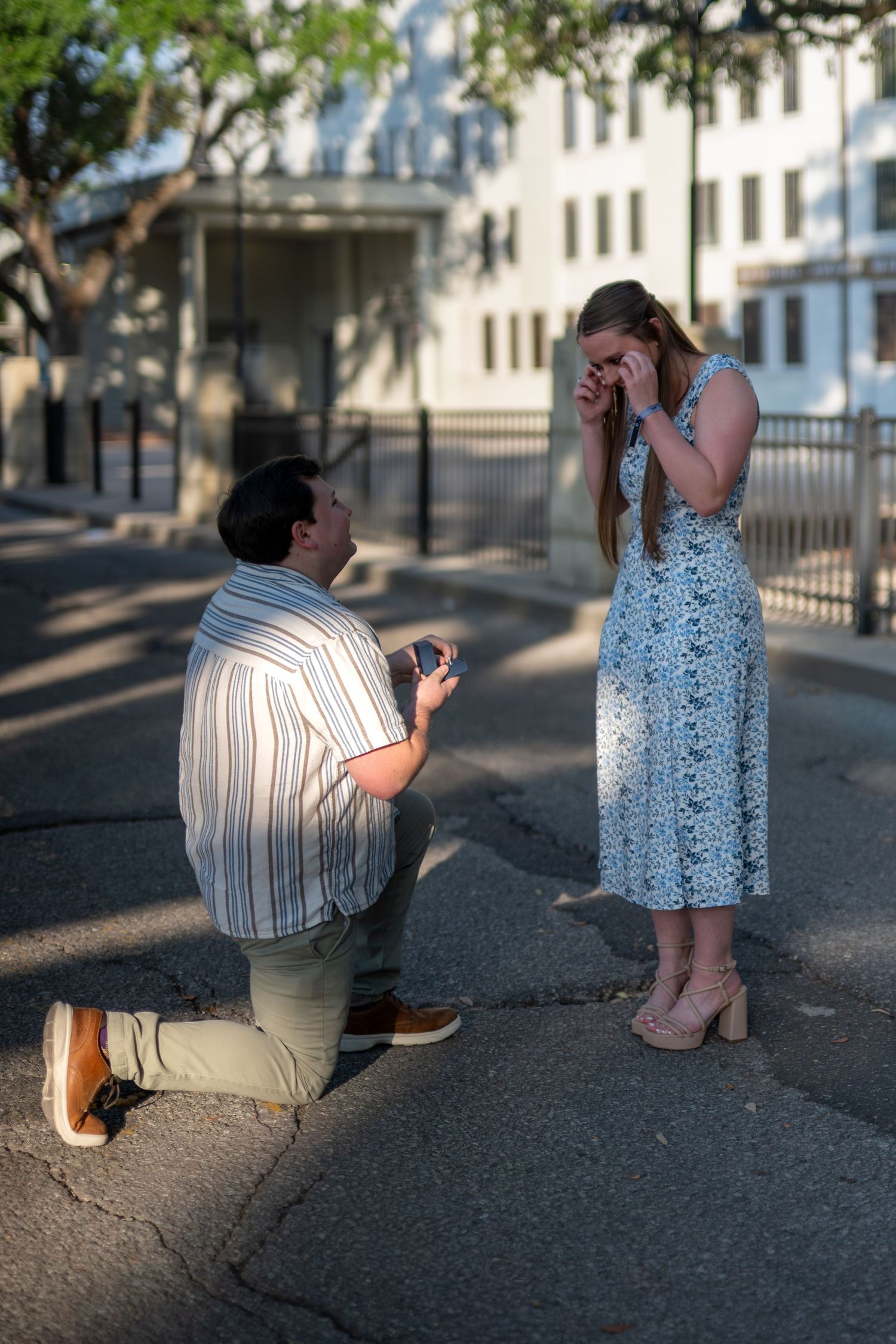 Man kneeling on one knee proposes to a woman covering her face with her hands in a sunlit street near a white building.