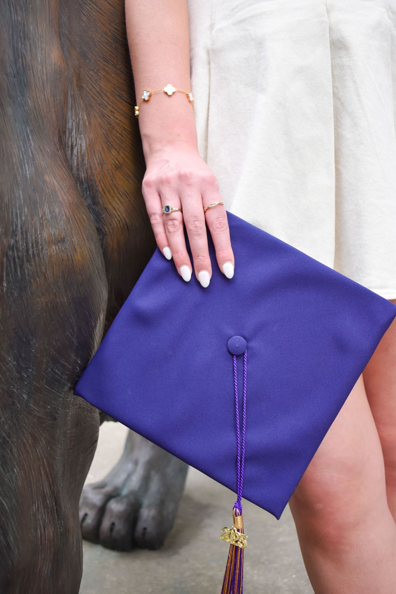 Graduate holding blue mortarboard cap with tassel beside bronze statue, wearing white dress.