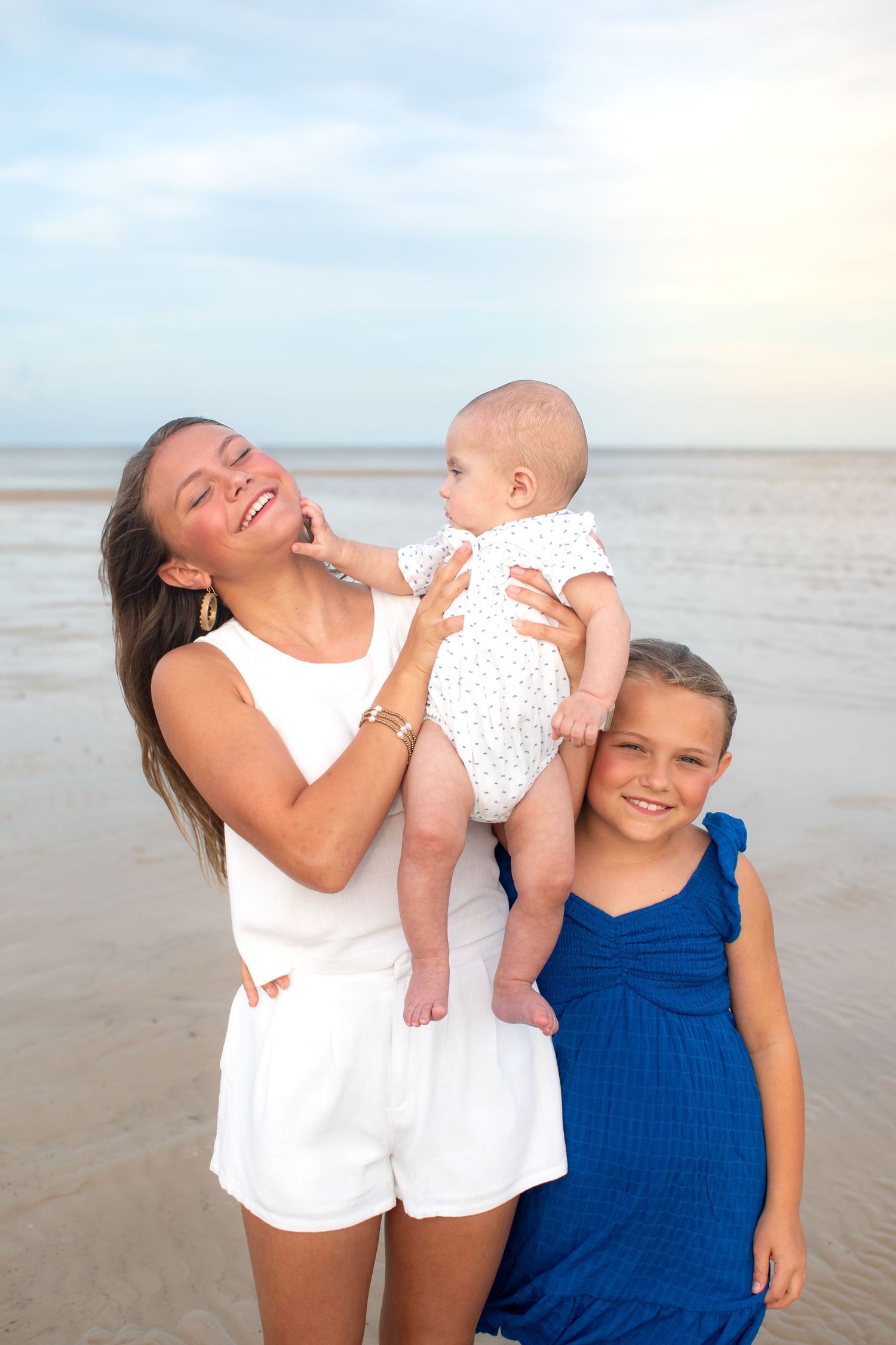 Joyful mother holds baby while older daughter in blue dress smiles on a sandy beach.