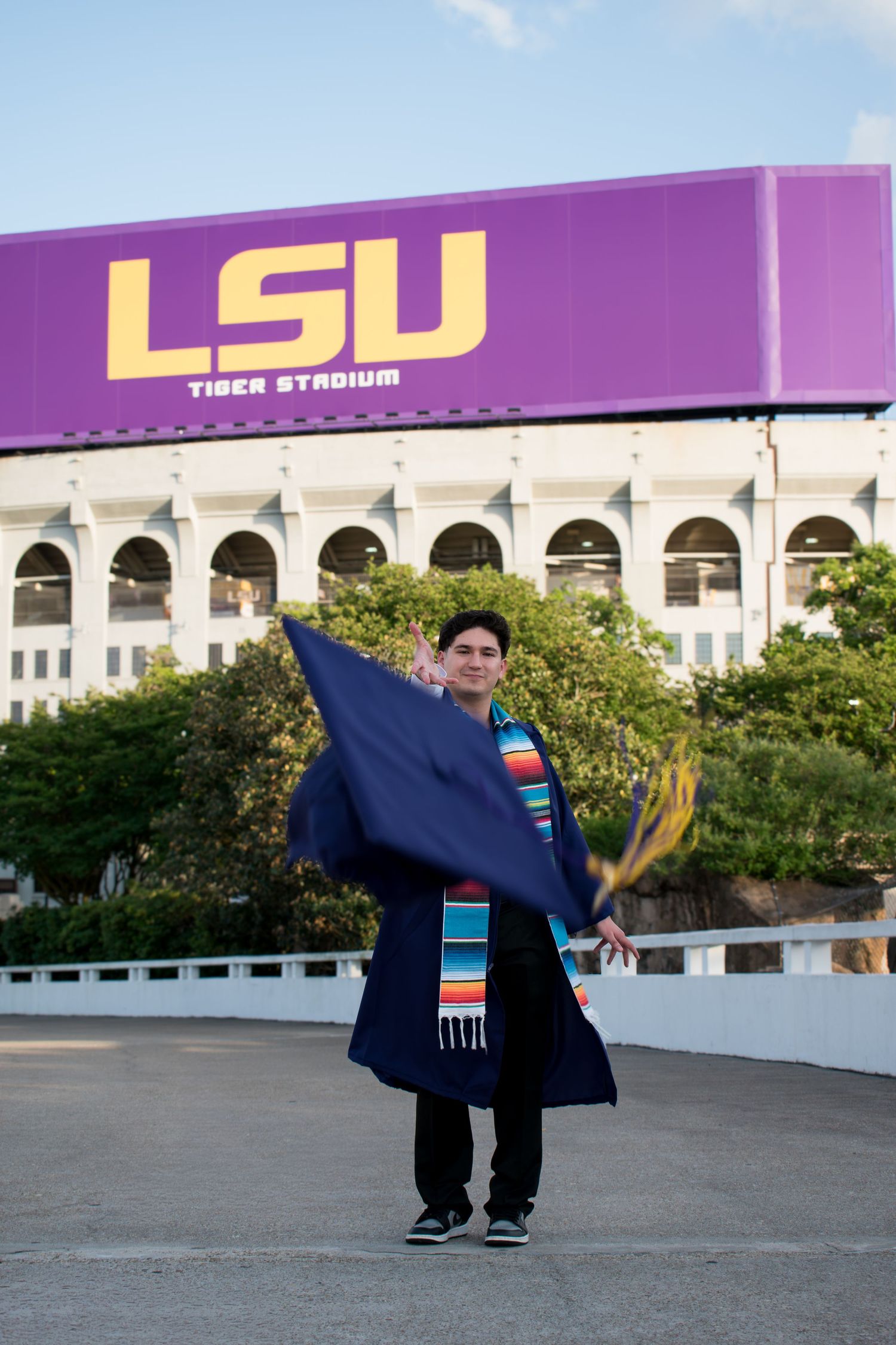 LSU graduate twirling blue graduation gown in front of Tiger Stadium's purple and gold sign.