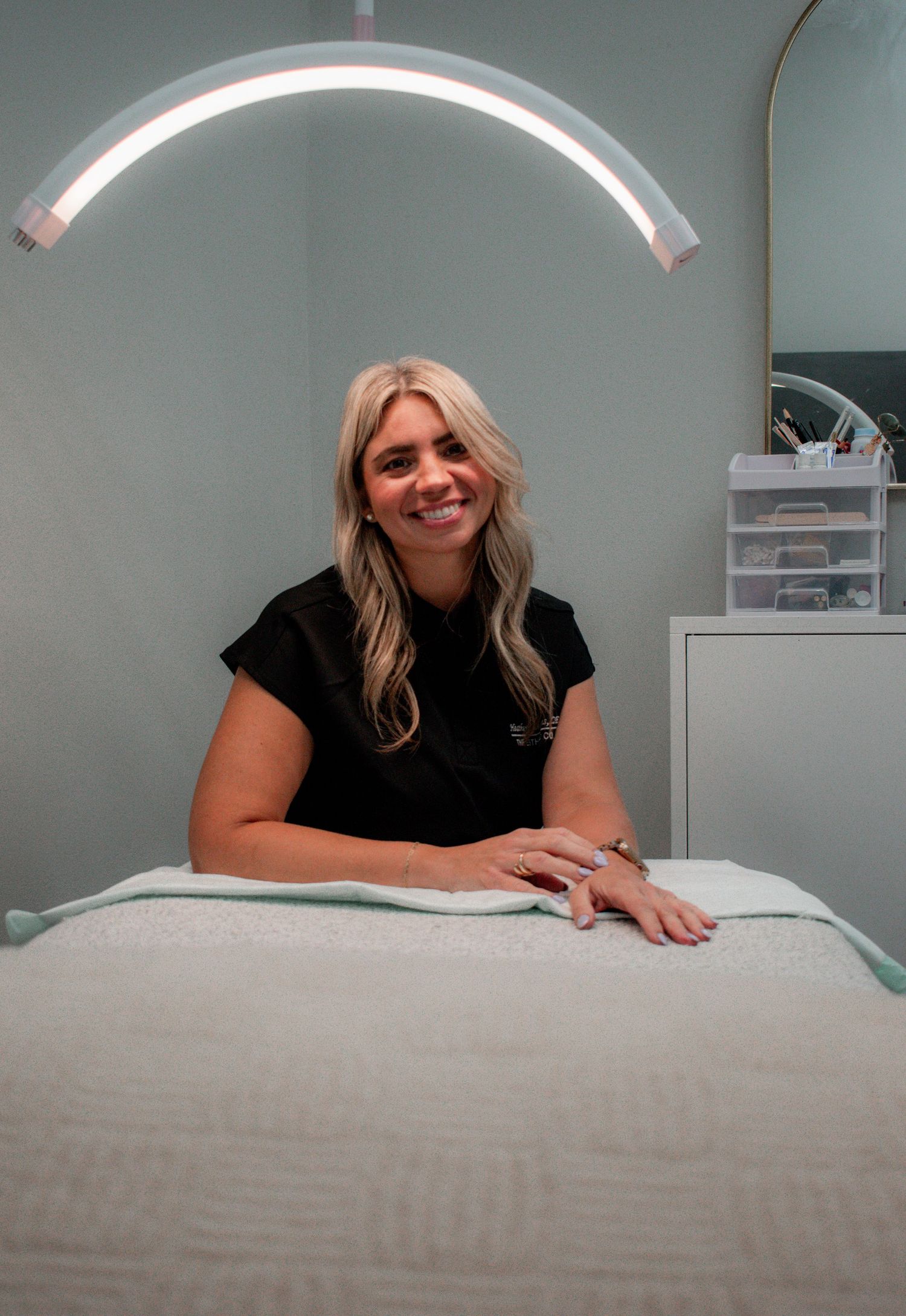 A blonde esthetician in black scrubs sits at a treatment table under an arch light in a spa room.