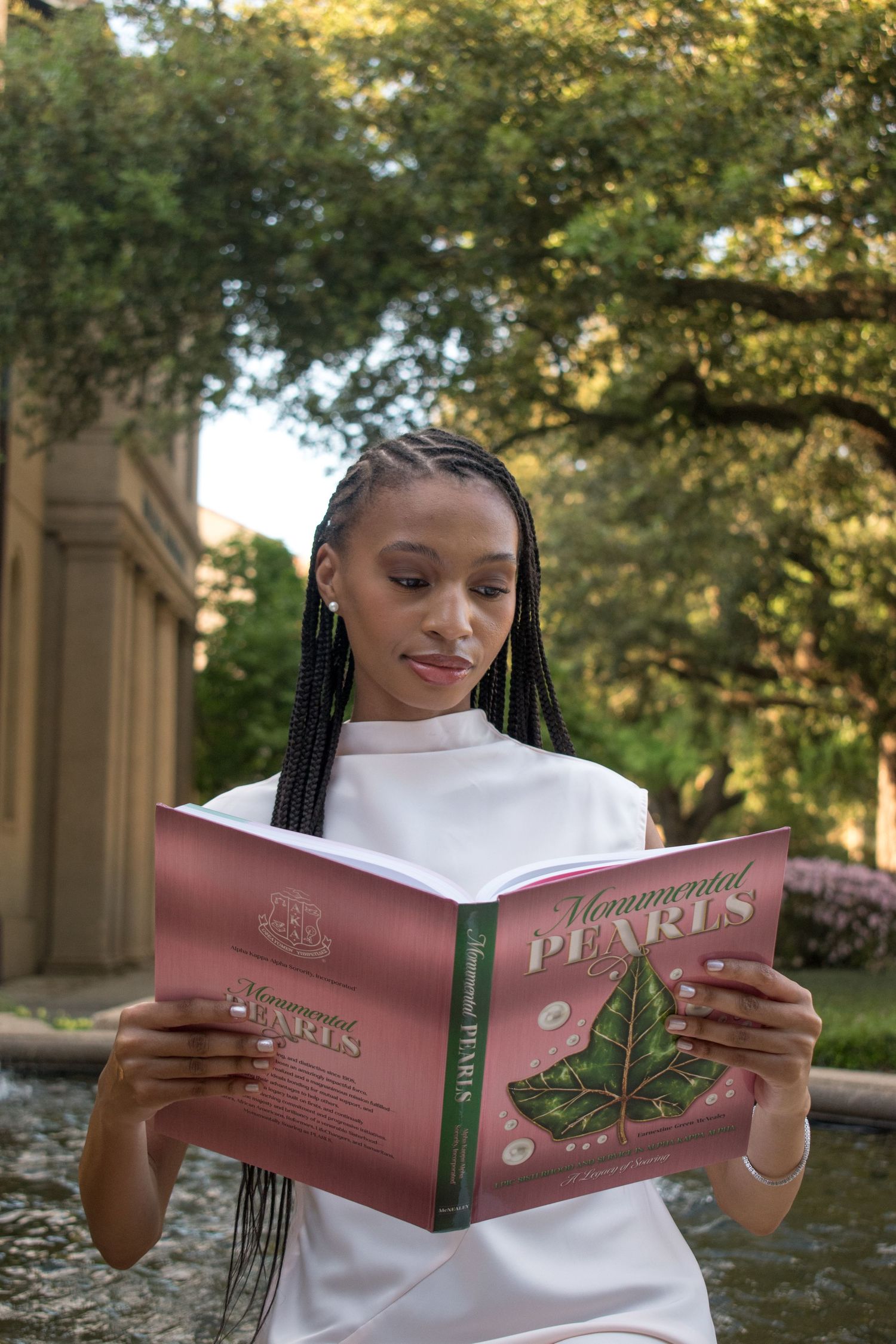 Young Black woman with braids in white dress reading Monumental Pearls book outdoors.