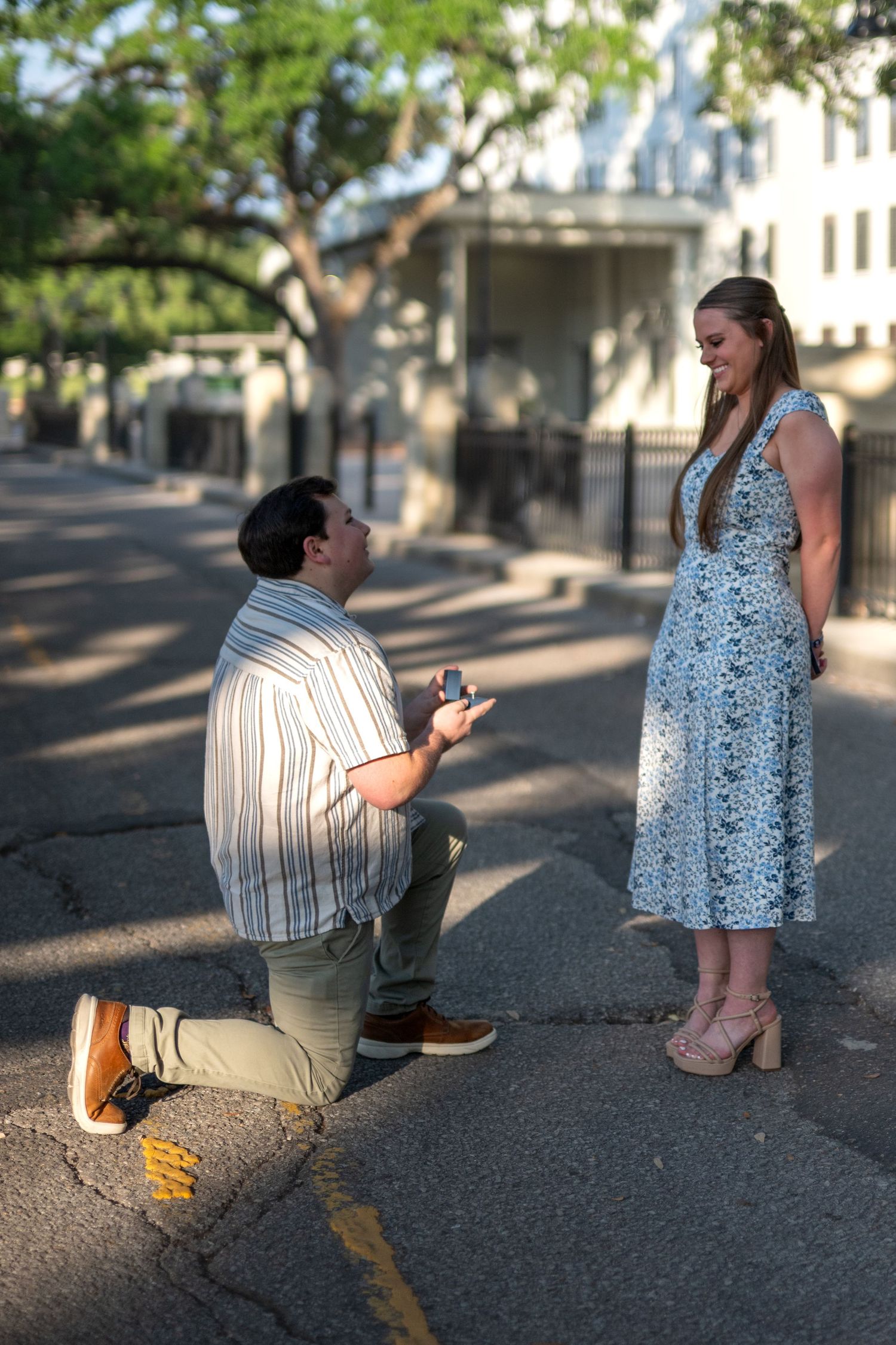 Man kneeling on one knee proposing to a smiling woman in a floral dress on a tree-lined street.