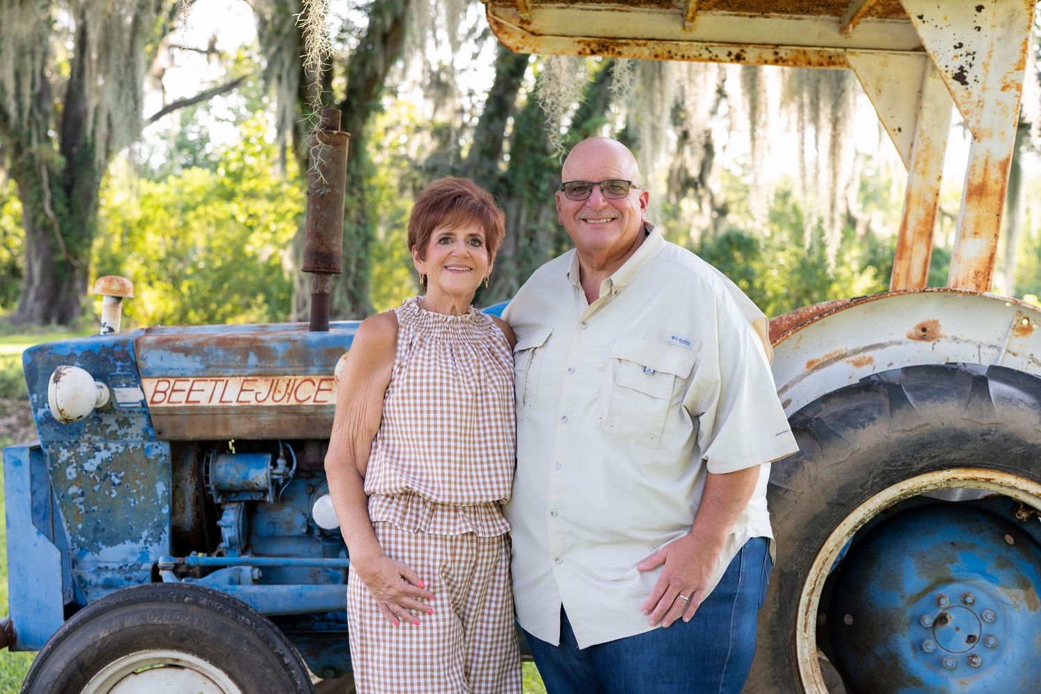 Smiling couple standing arm in arm beside a vintage blue tractor with oak trees draped in Spanish moss in the background.