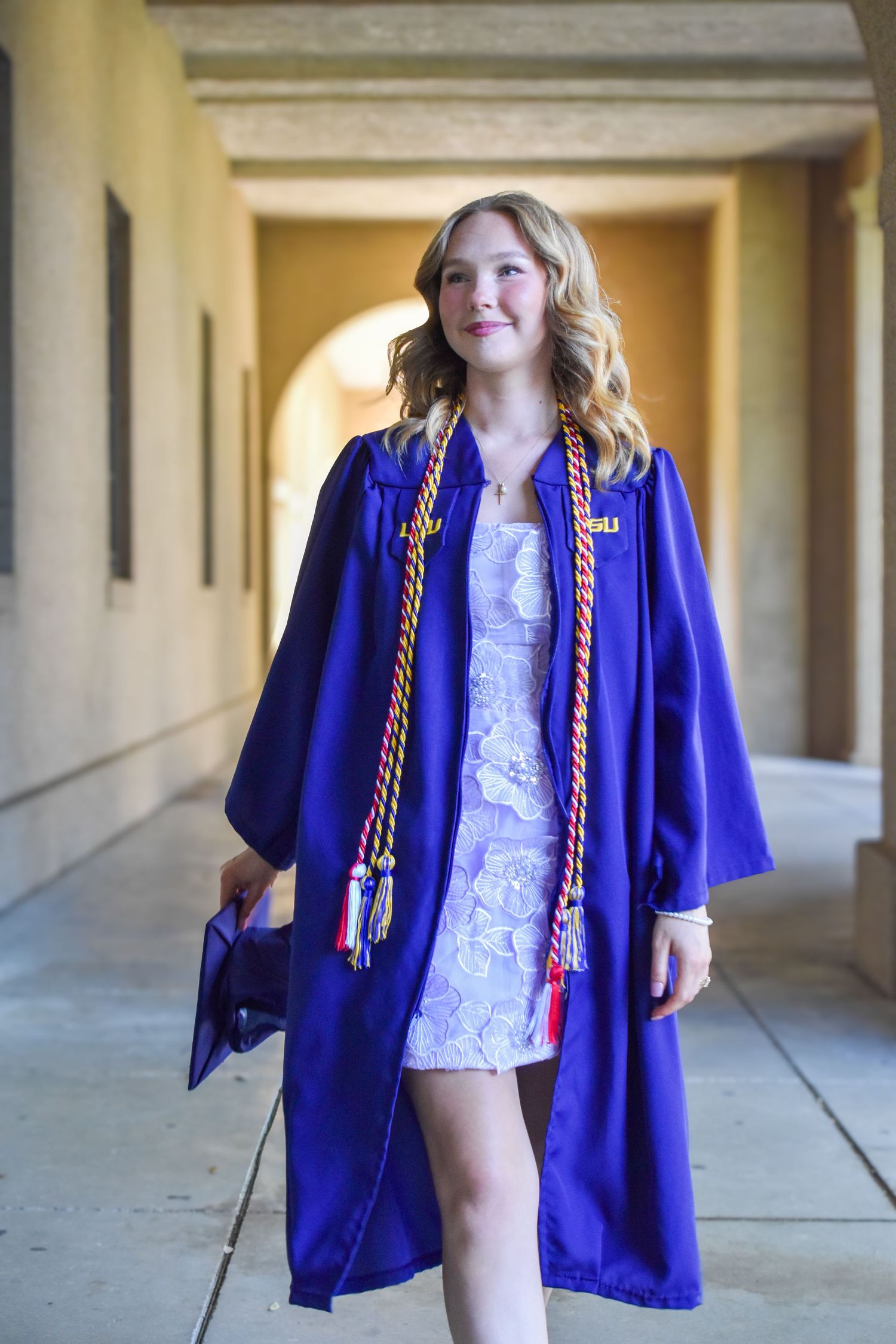 Female graduate walking in blue cap and gown with honor cords in university hallway.