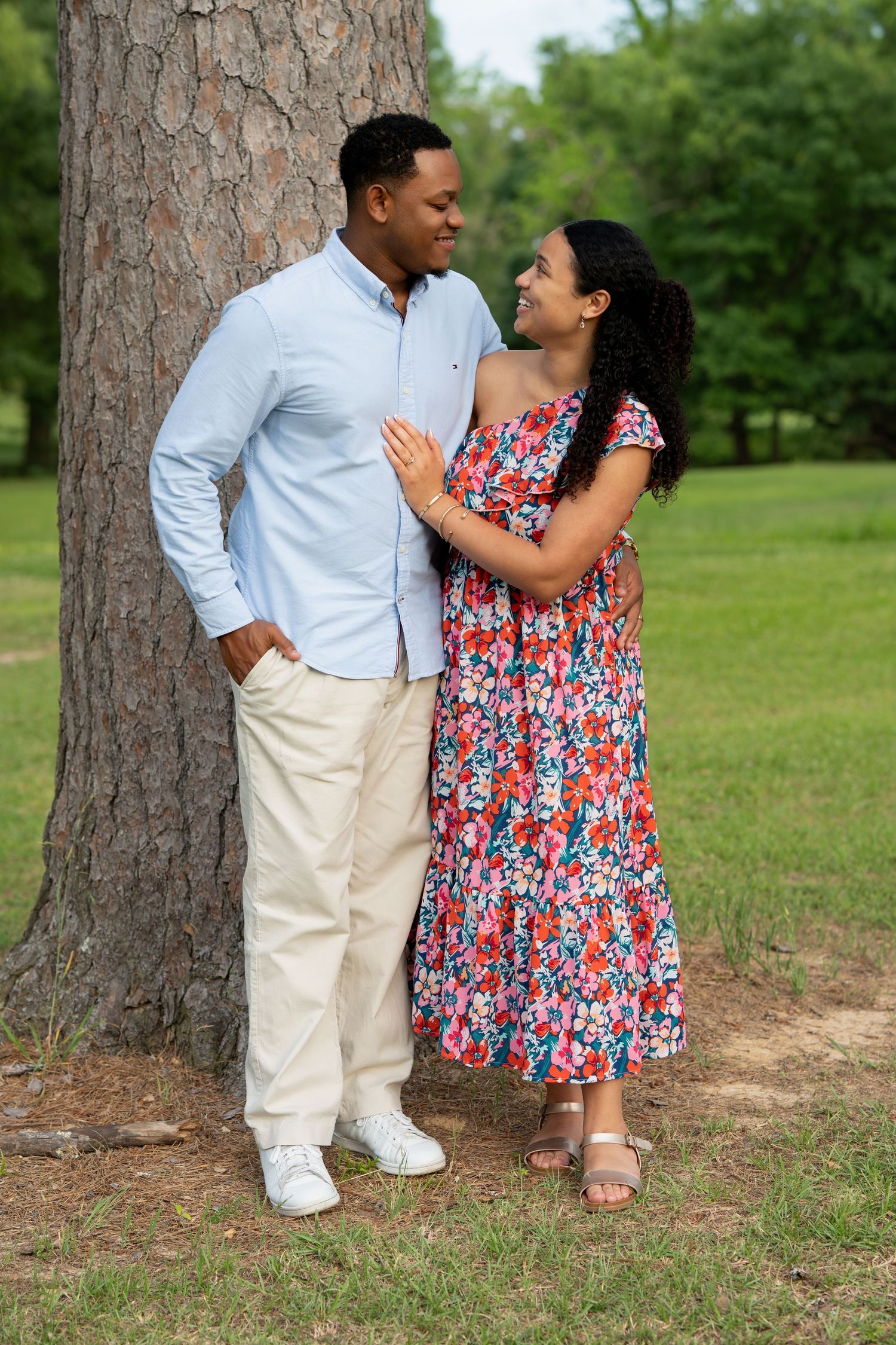 A couple shares a romantic moment leaning against a tree in a lush green park, dressed in casual summer attire.