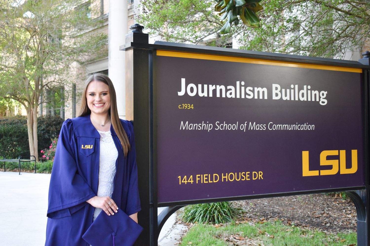 Female LSU graduate in blue gown posing beside Manship School of Mass Communication sign.