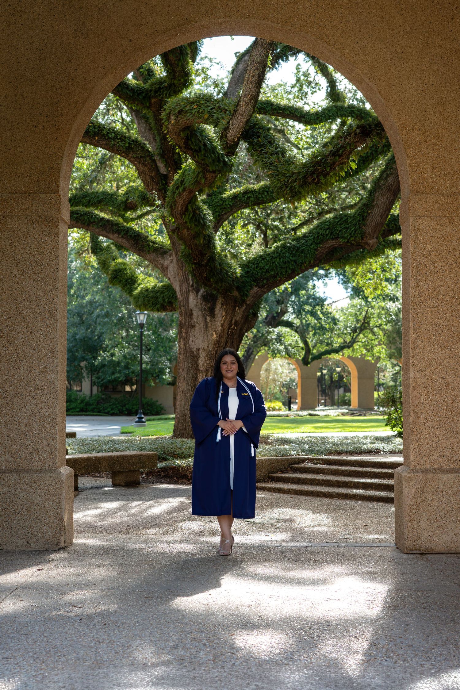 Graduate in blue cap and gown stands under stone arch framed by a large moss-covered oak tree on campus.