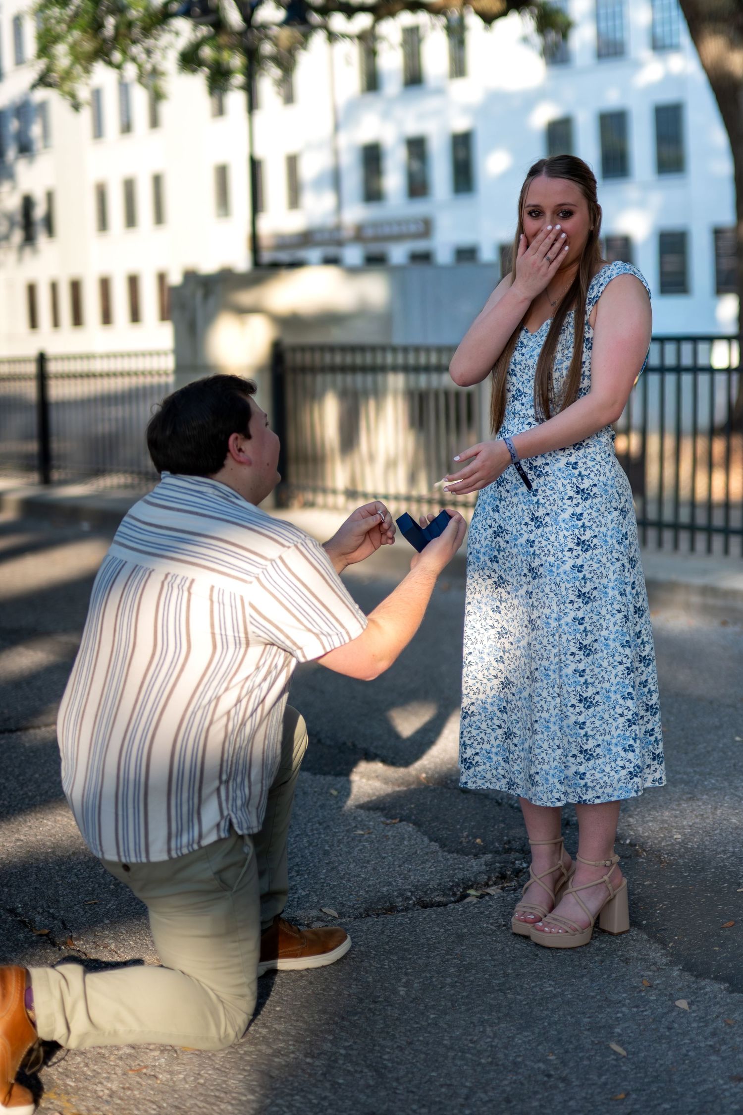 Man kneeling on one knee proposing to surprised woman in floral dress on sunny urban sidewalk.