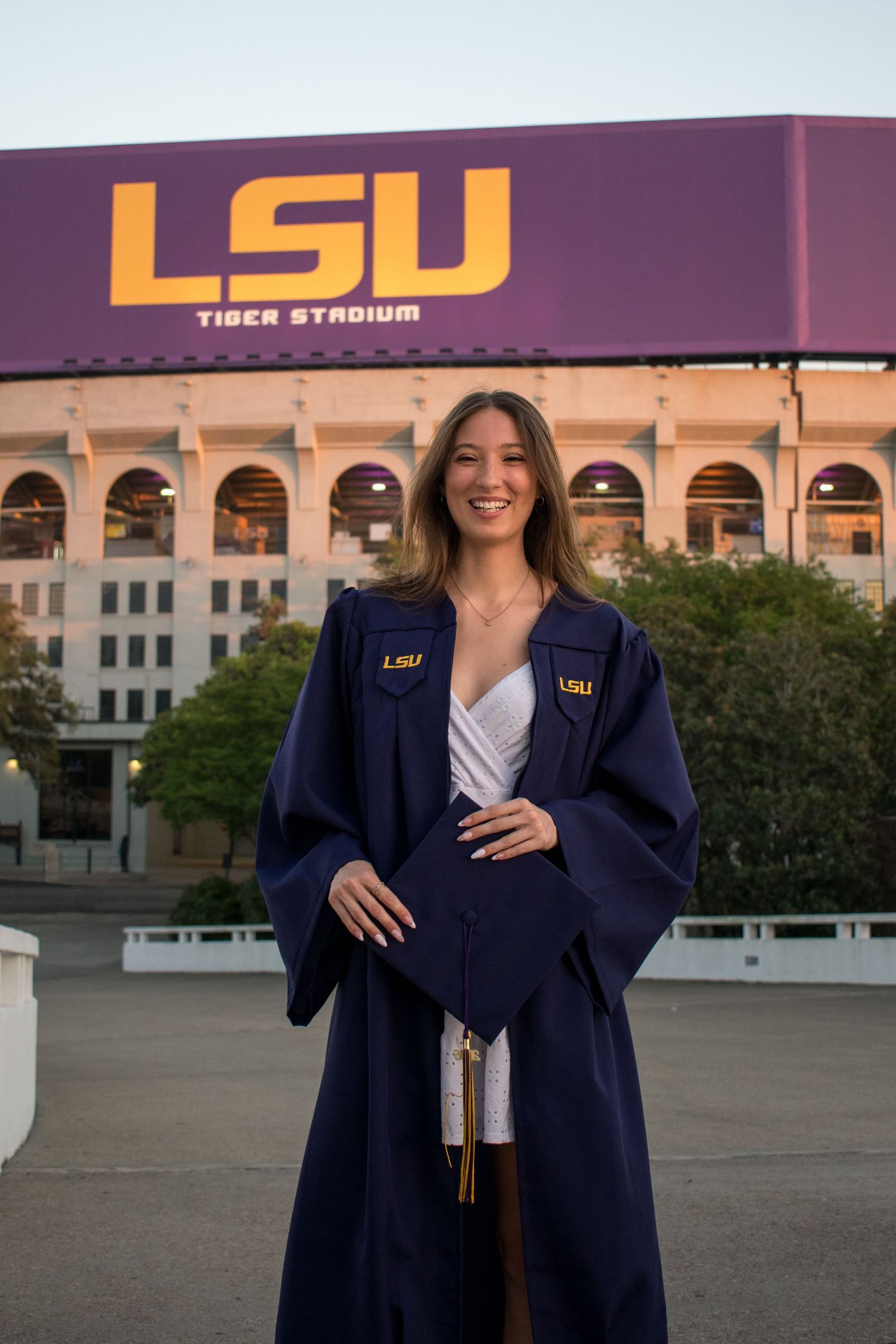 Female graduate in LSU navy gown smiling in front of illuminated Tiger Stadium at night.